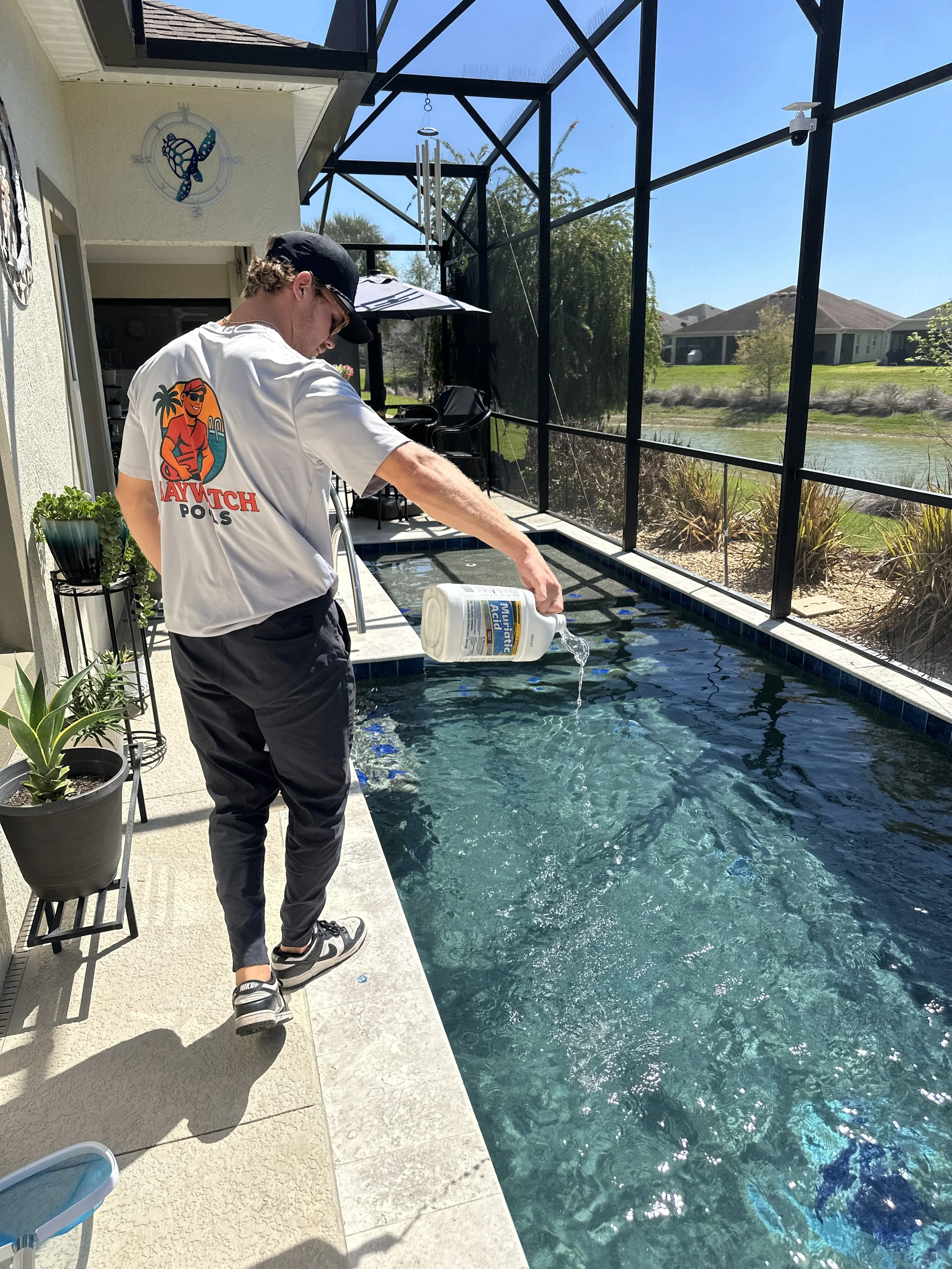 A young man is pouring a bottle of water into a swimming pool on a sunny day. He is wearing a white t-shirt with a colorful graphic, black pants, and sneakers. The pool area is enclosed with a screened structure, and there are plants and houses visible outside.