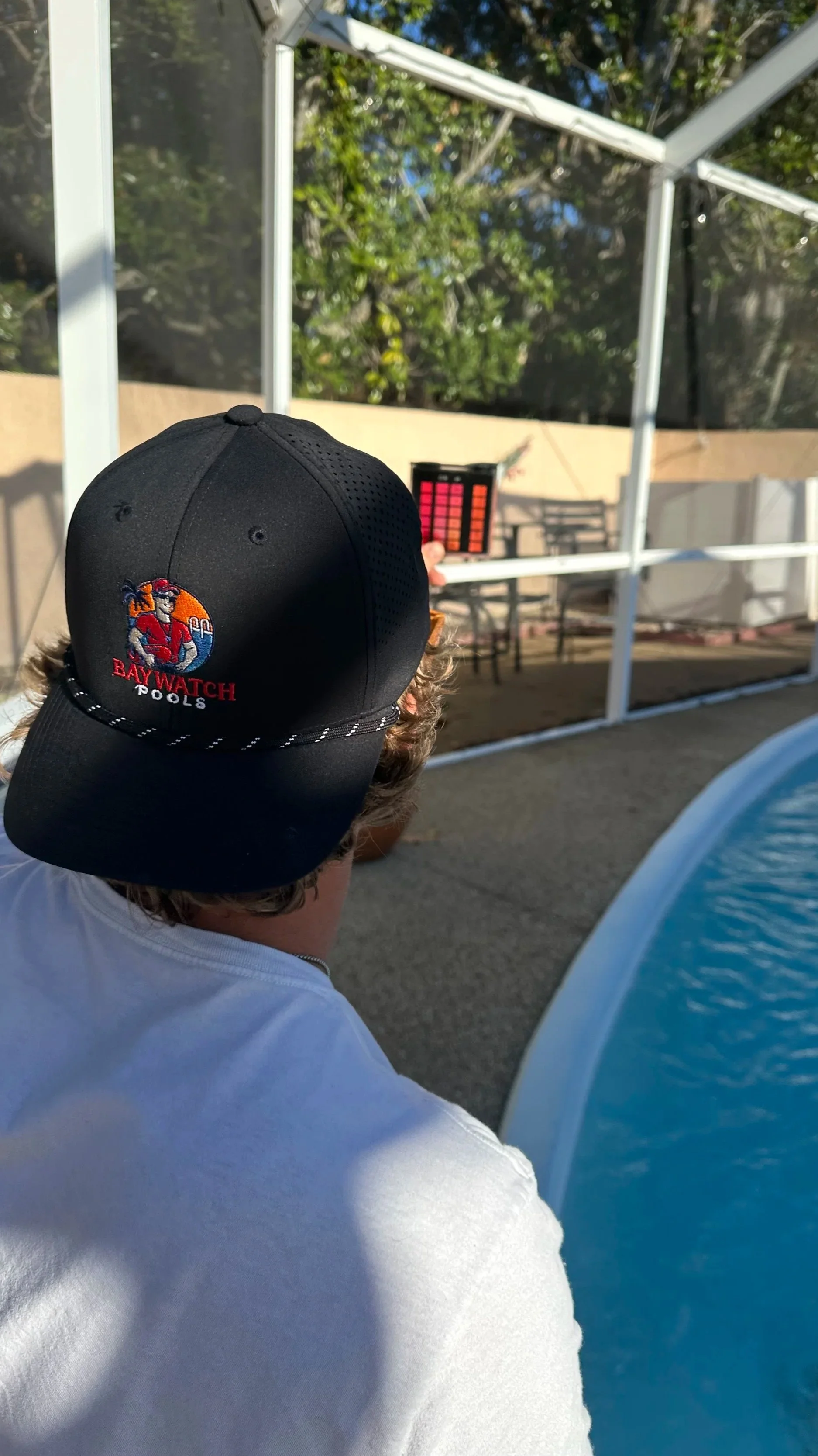 Person wearing a black baseball cap with a 'Baywatch Pools' logo, facing away, at an outdoor pool area with a person in the background holding a color palette, and pool with blue water in the foreground.