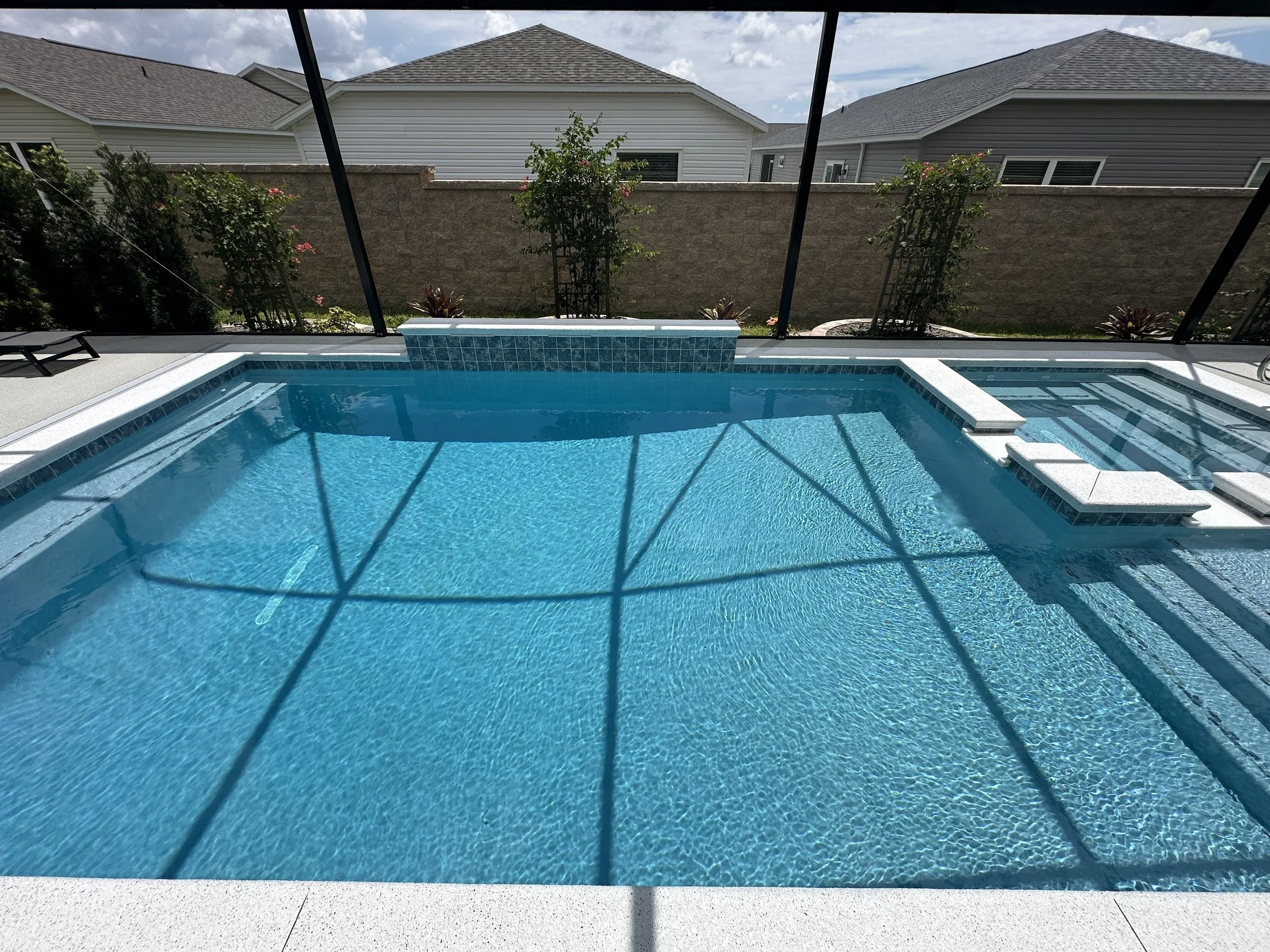 Backyard swimming pool with a spa and a screened enclosure, surrounded by a concrete deck and a garden with trees and shrubs, with neighboring houses and cloudy sky in the background.