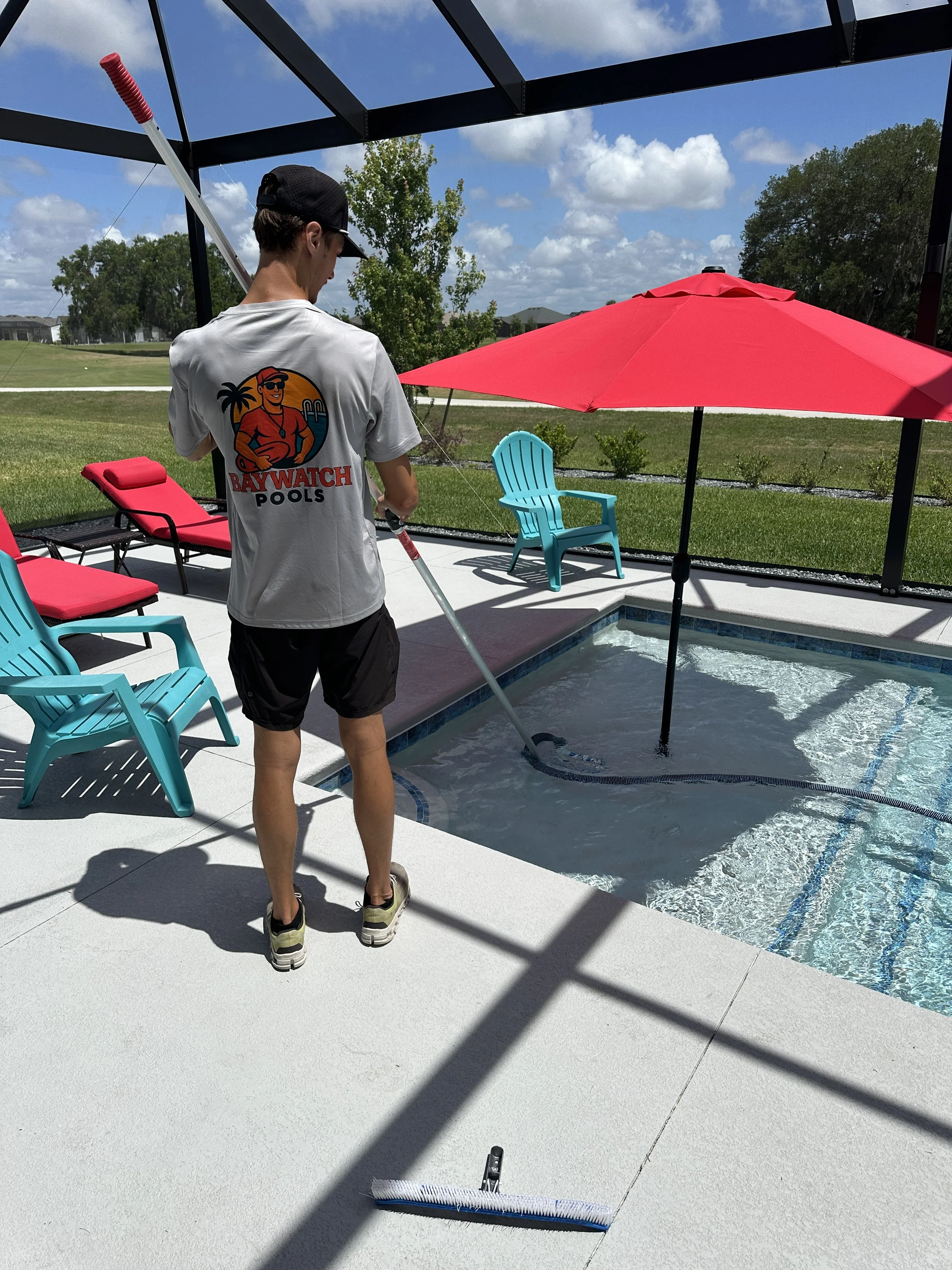 Person standing by a pool cleaning equipment with a pool cover in the background, outdoor setting with lounge chairs, an umbrella, and a blue sky with clouds.