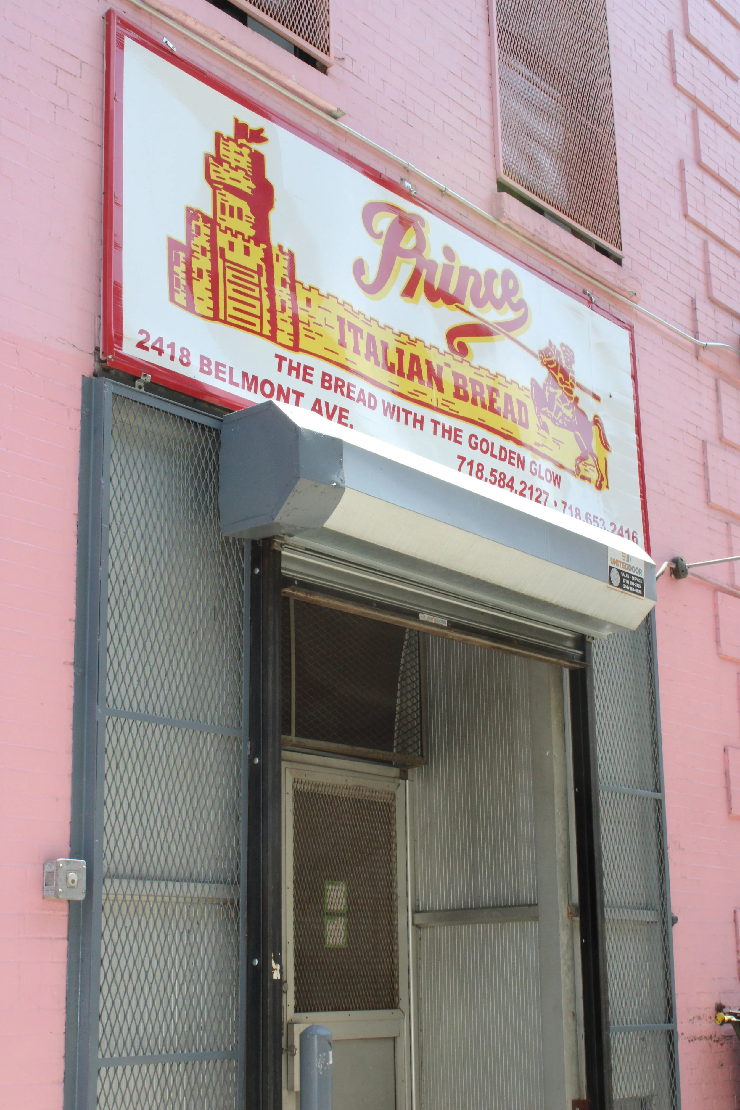 A storefront with a sign for Prince Italian Bread, showing the address 2418 Belmont Ave and contact numbers, with a pink brick building and an open metal security gate.