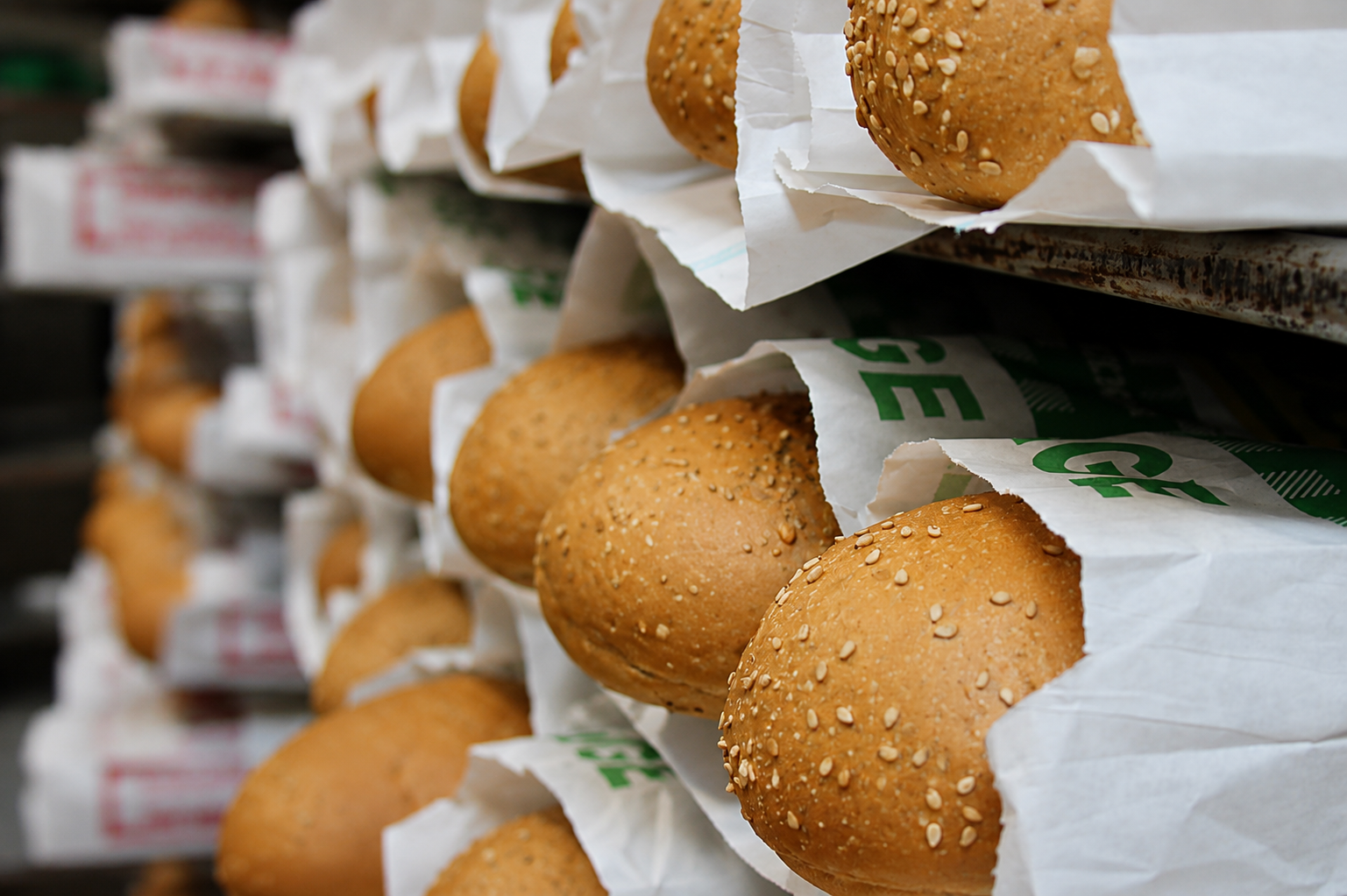 Multiple sesame seed hamburger buns wrapped in white bakery paper, stacked on bakery shelves.