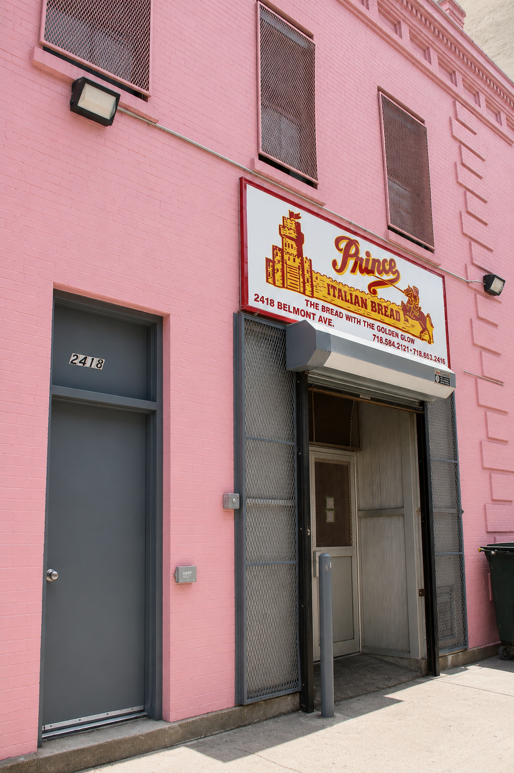 The entrance of a pink building with a sign reading "Prince Italian Bread" and a door numbered 2418, located on Belmont Avenue, displaying contact numbers."