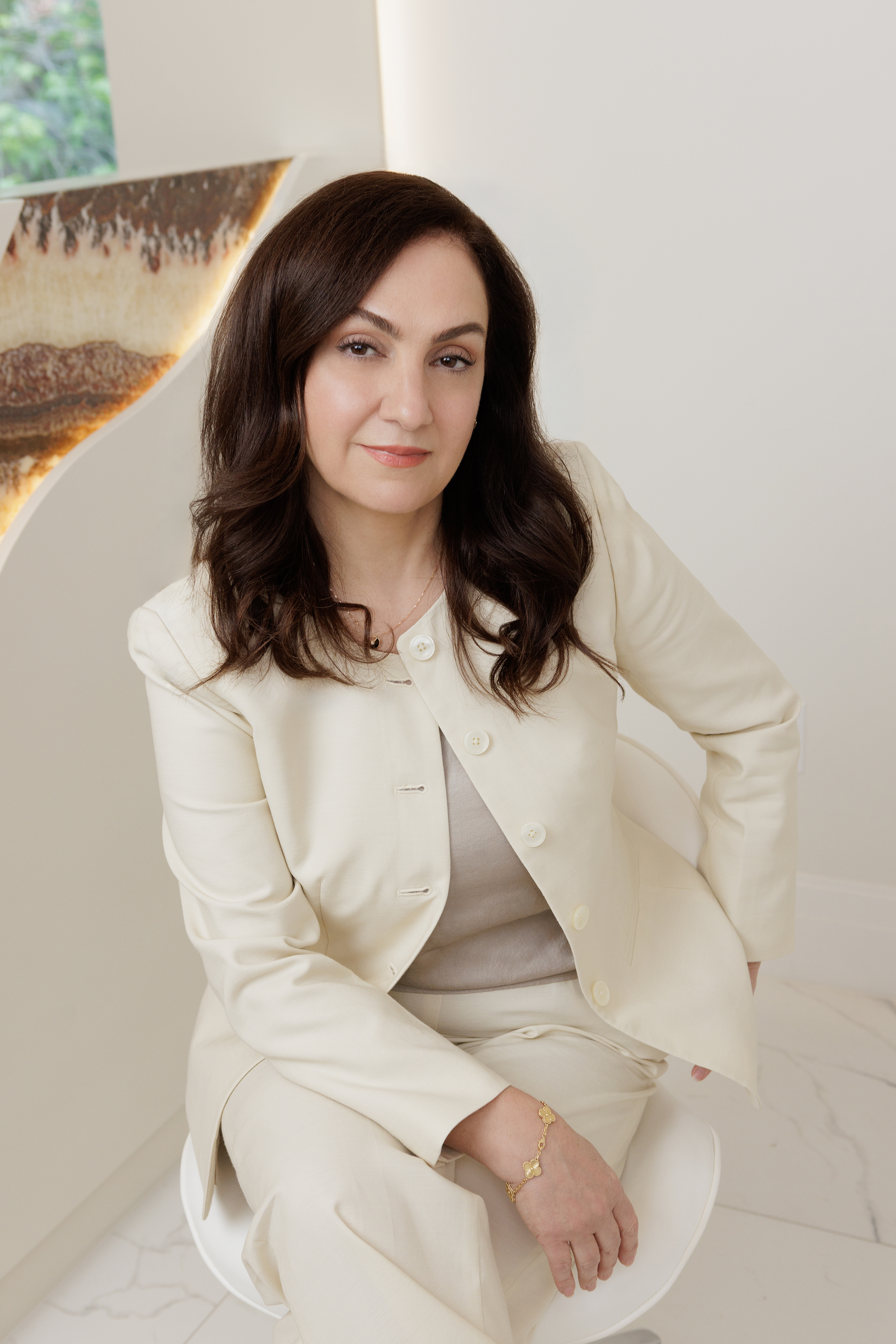 A woman with shoulder-length dark hair and light skin, wearing a cream-colored blazer, sitting on a white chair in a modern, minimalist room with white walls and marble flooring. She has a calm expression with a slight smile.