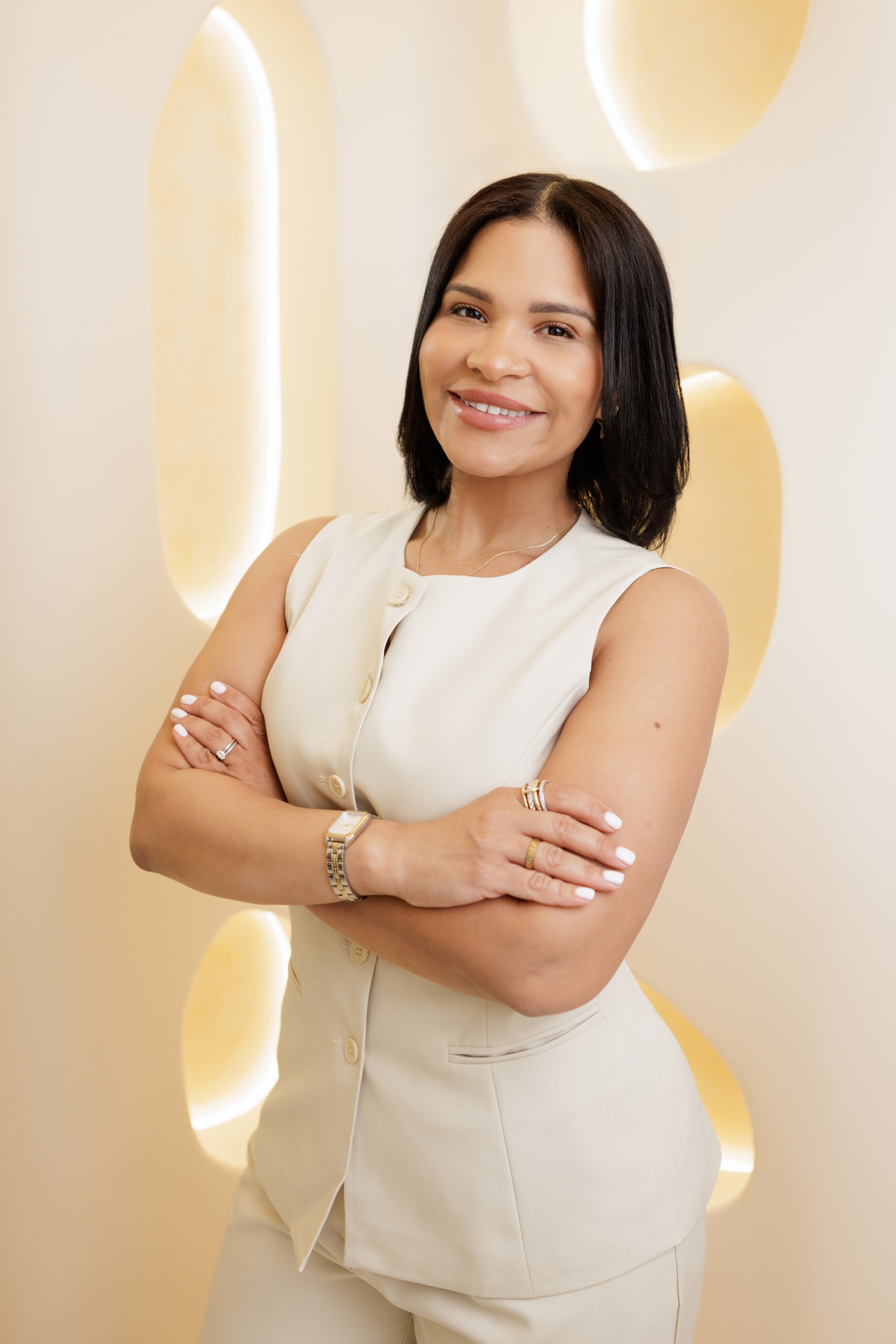 A woman with shoulder-length dark hair smiling and standing with arms crossed, wearing a sleeveless beige blazer and matching pants, in front of a beige wall with circular cutouts illuminated with light.