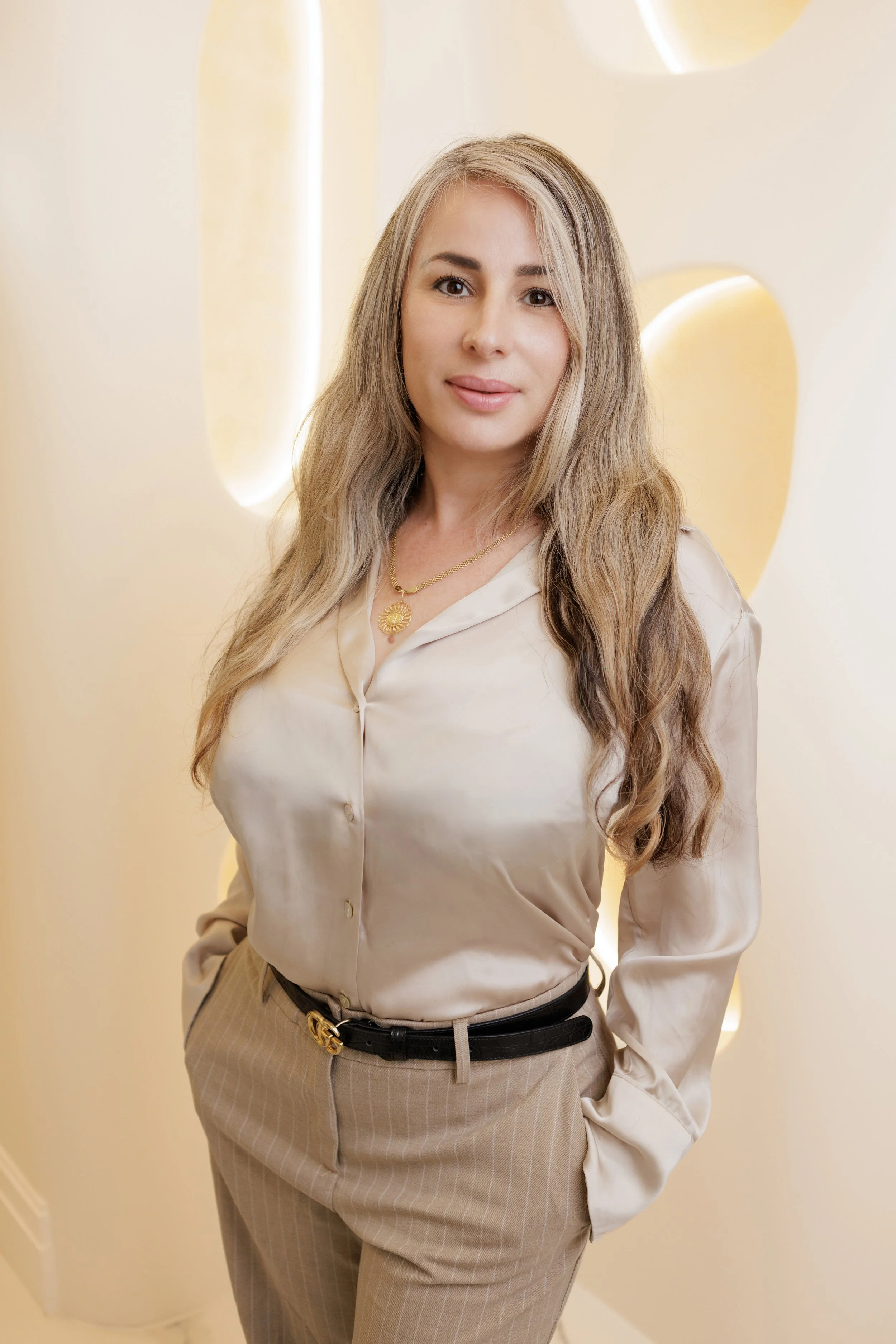 A woman with long, wavy blonde hair wearing a beige satin blouse and striped tan pants with a black belt, standing against a cream-colored wall with rounded decorative elements.