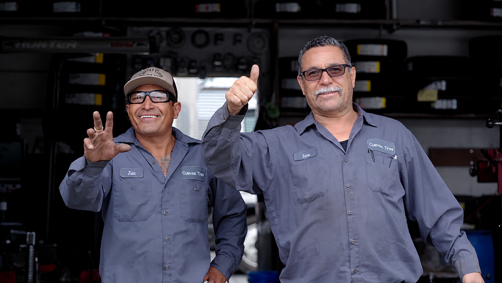 Two smiling mechanics wearing gray uniforms and glasses, standing in a garage, making gestures; one hand showing a peace sign and the other with a thumbs up.