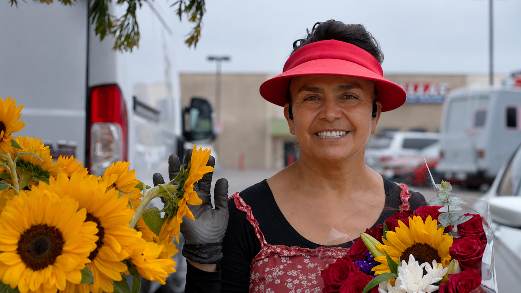 A woman wearing a red hat, black gloves, and a black and red apron, smiling while holding a bouquet of sunflowers and red roses, standing outdoors in a parking lot with vans and a building in the background.