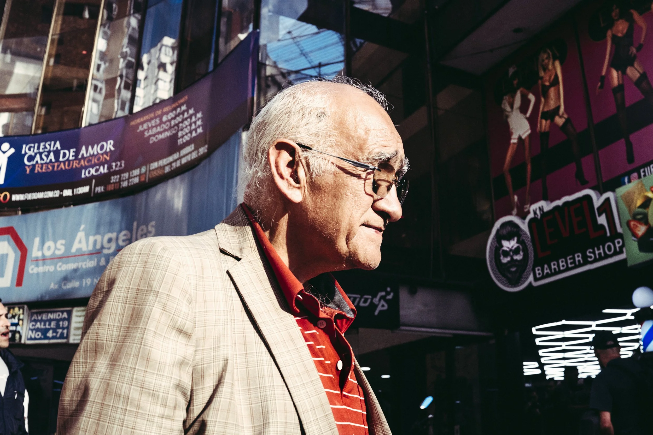 Un hombre mayor con gafas y chaqueta a cuadros caminando por una calle concurrida en Los Ángeles, con anuncios y carteles publicitarios en el fondo.