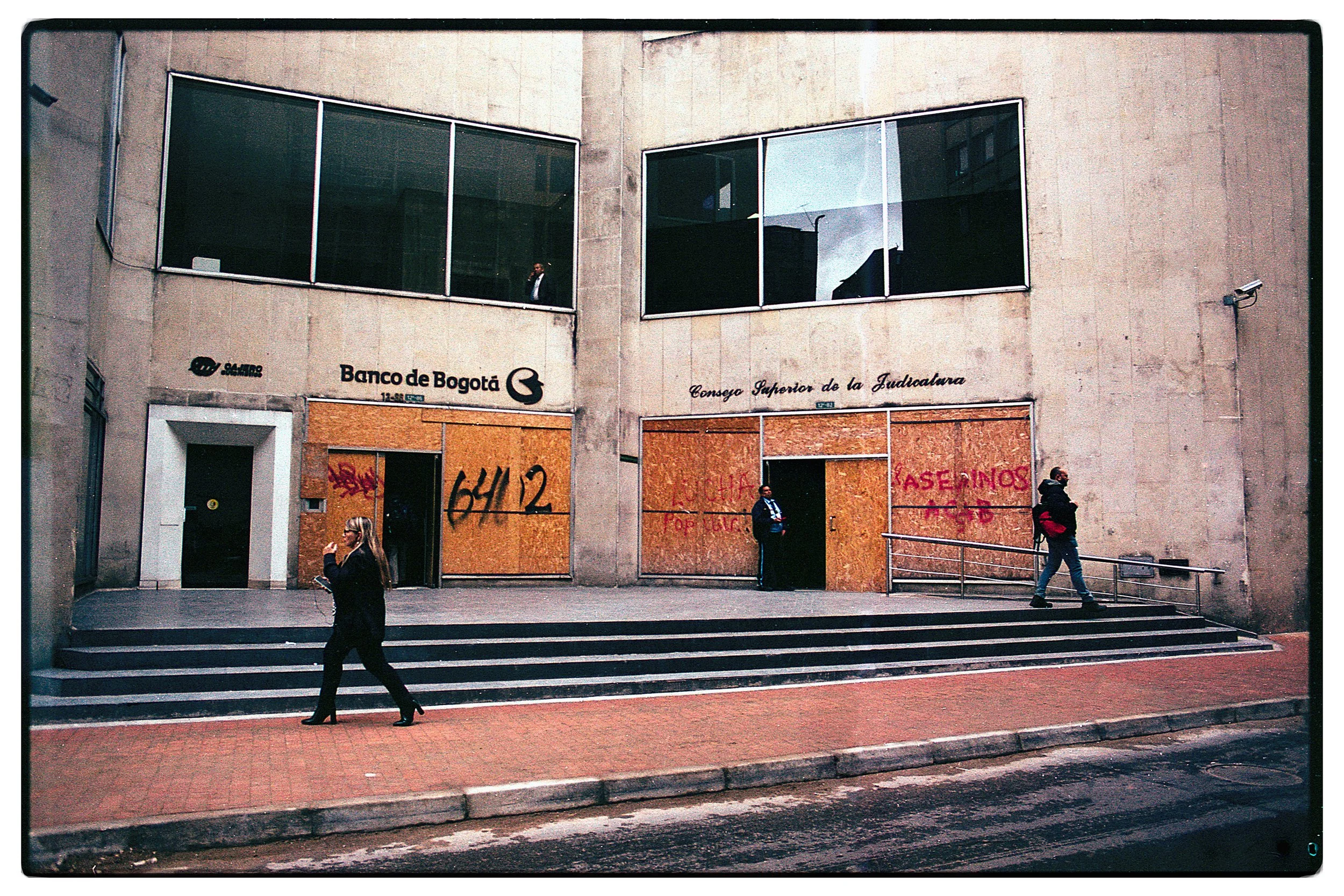 Edificio con ventanas de vidrio y paredes de concreto. Personas caminando en la acera en frente del edificio, algunos con mascarillas y otros sin ellas.