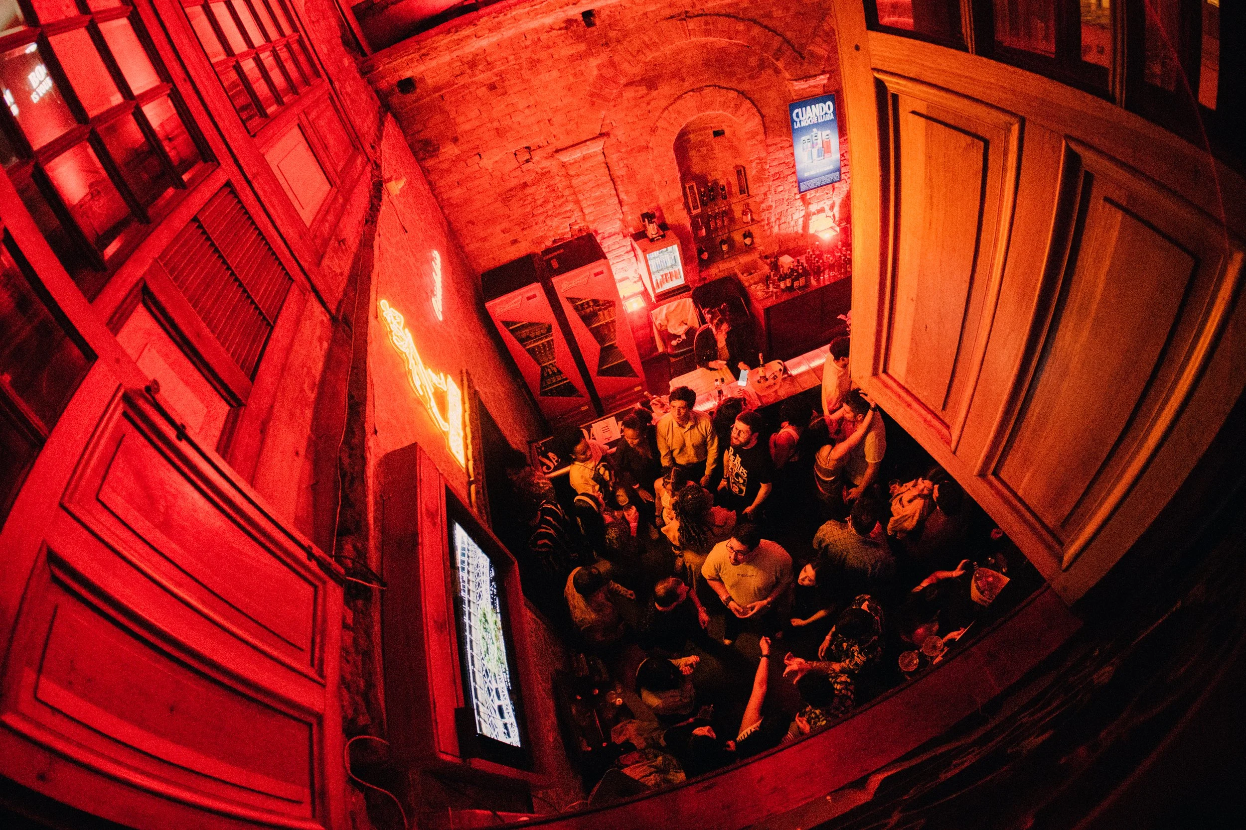 Interior de un bar o restaurante con paredes de ladrillo, neon rojo y carteles, con varias personas conversando y tomando bebidas.