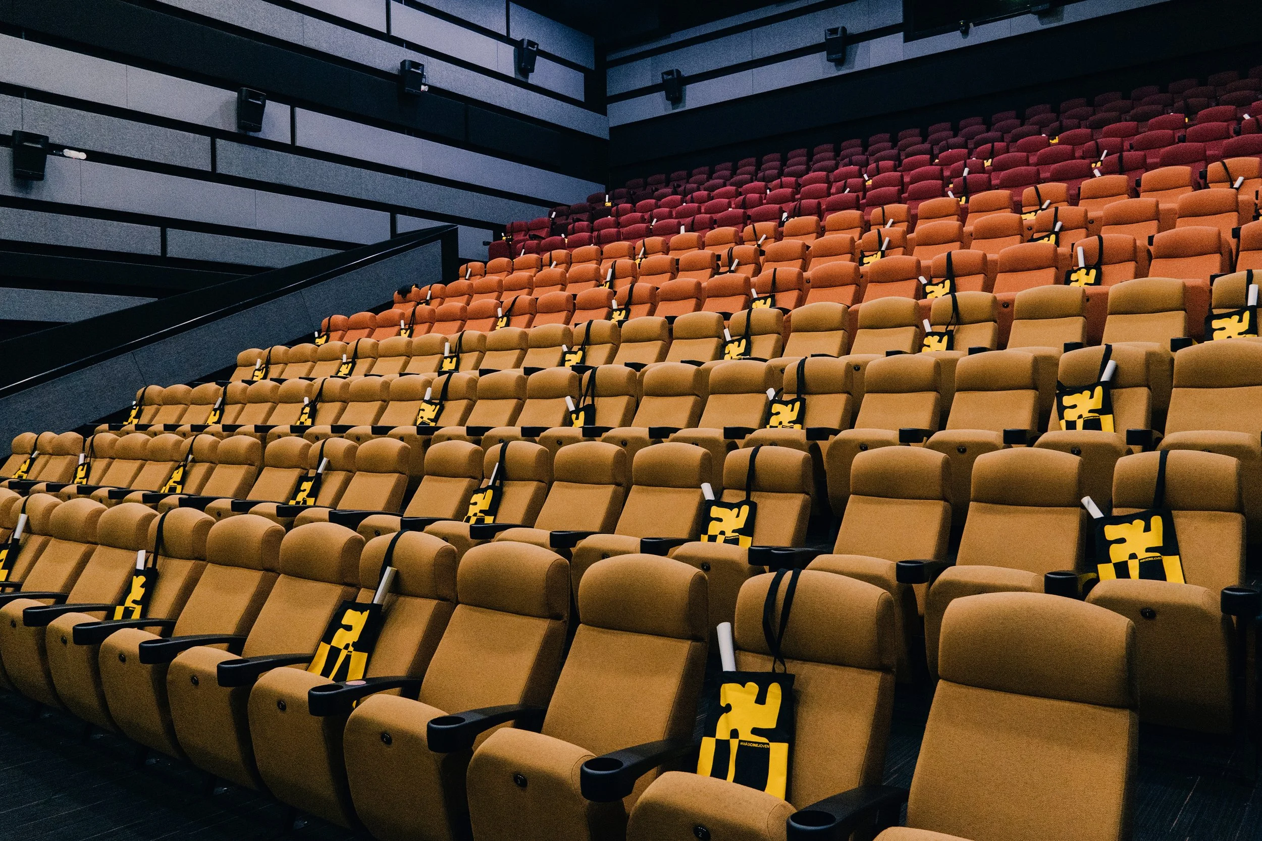 Sala de cine con asientos en tonos beige y naranja, algunos con mascarillas de seguridad colgadas.
