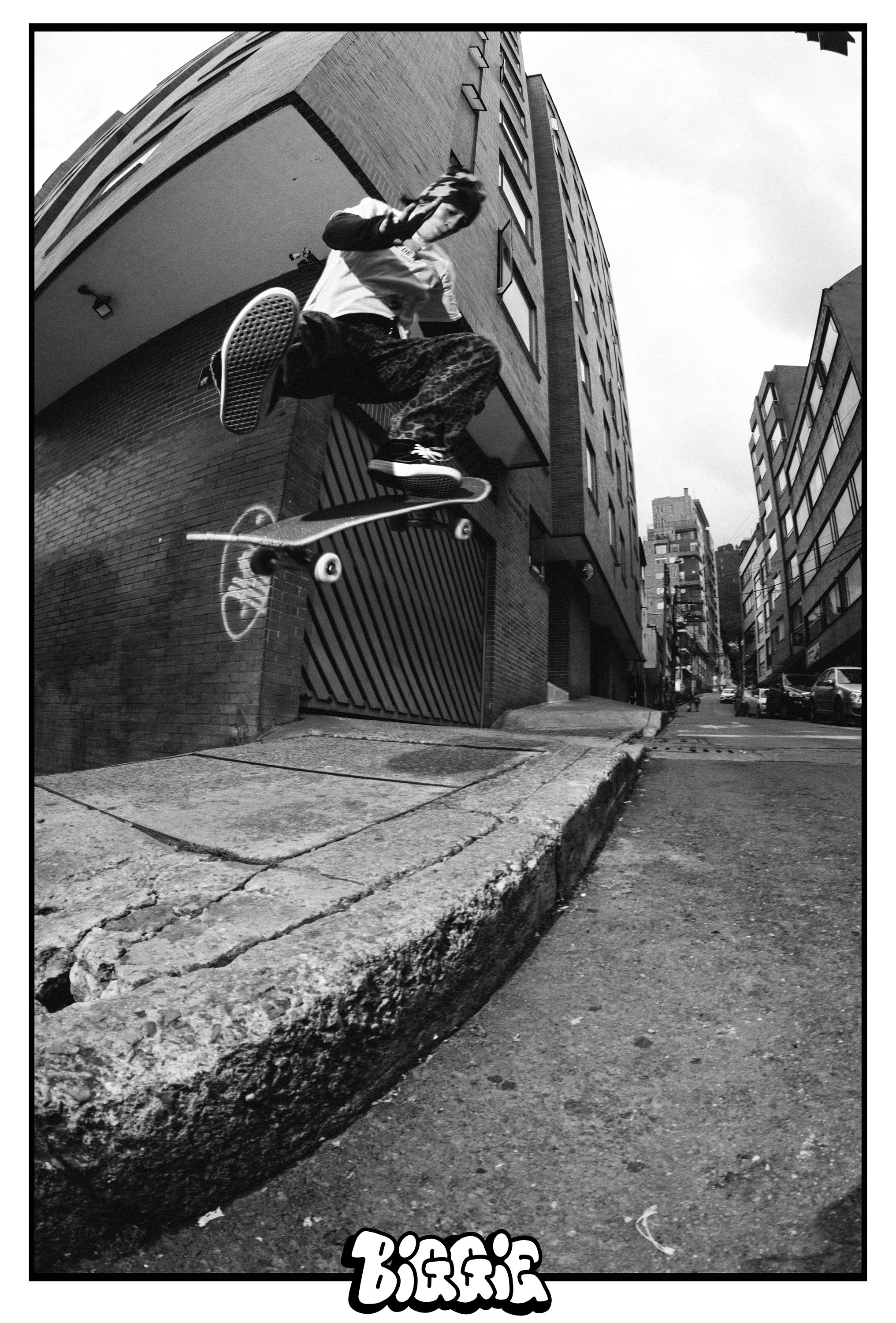 Chico haciendo skate en la acera de una calle urbana, edificios de ladrillo y calles con autos en el fondo, en blanco y negro.