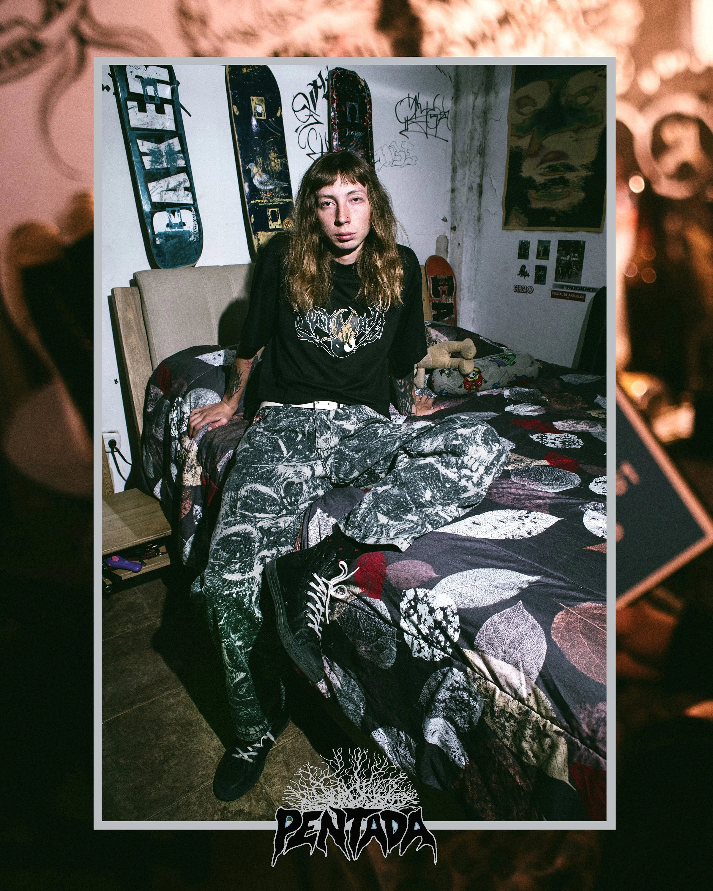 Joven con cabello largo y oscuro, usa camiseta negra con diseño y pantalones con patrón. Está en una habitación adornada con graffiti y pósters, sentado en una cama con edredón oscuro y rodeado de objetos diversos.