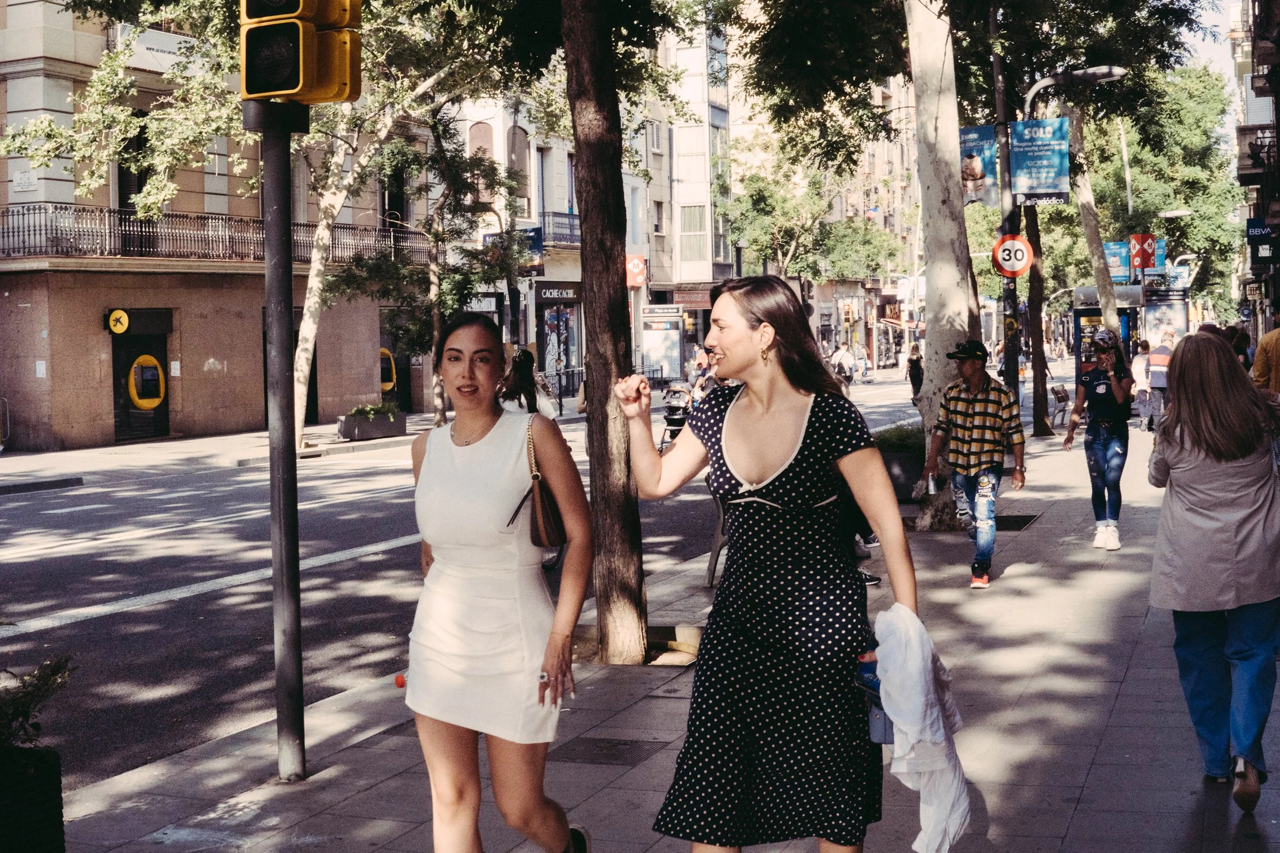 Dos mujeres caminando juntas en una calle concurrida, rodeadas de árboles, edificios y otros peatones.