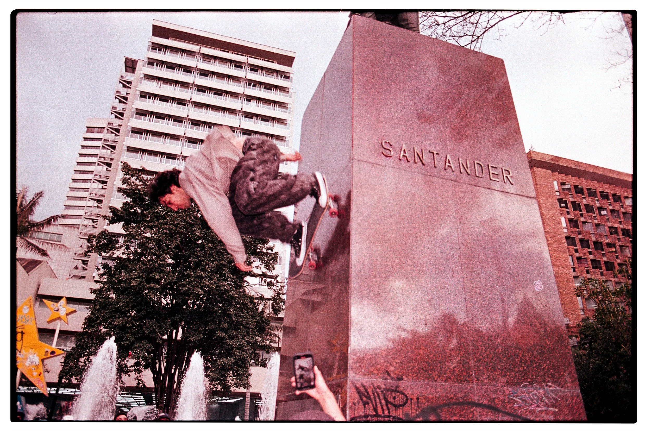 Una persona saltando sobre un monumento de piedra con la inscripción 'SANTANDER', en un entorno urbano con edificios altos, árboles y una fuente en primer plano.