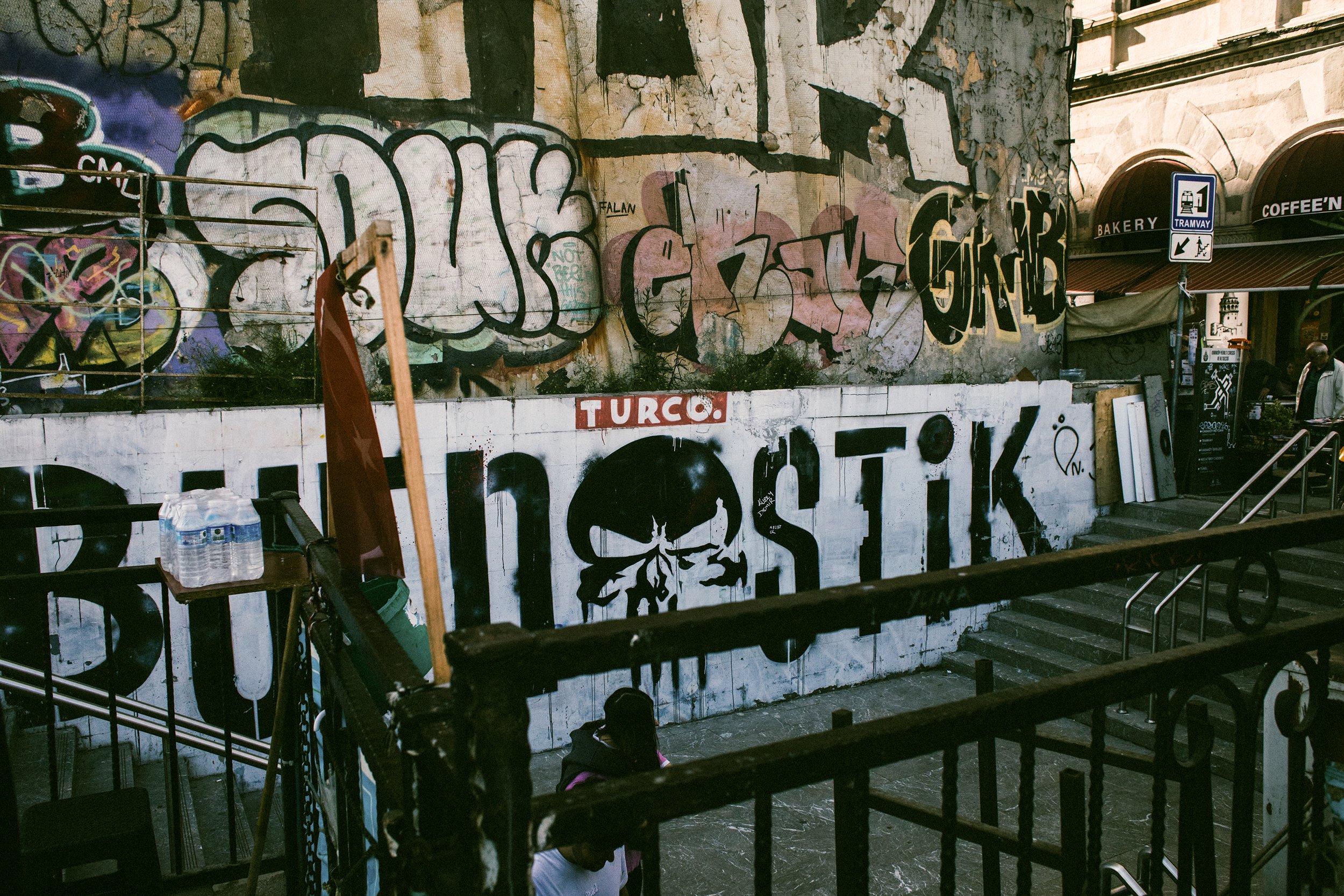 Muro con grafiti en un edificio deteriorado, con plantas creciendo en la base y una señal de tránsito que indica área de tranvía y salida de peatones, junto a unas escaleras y personas en la parte inferior.