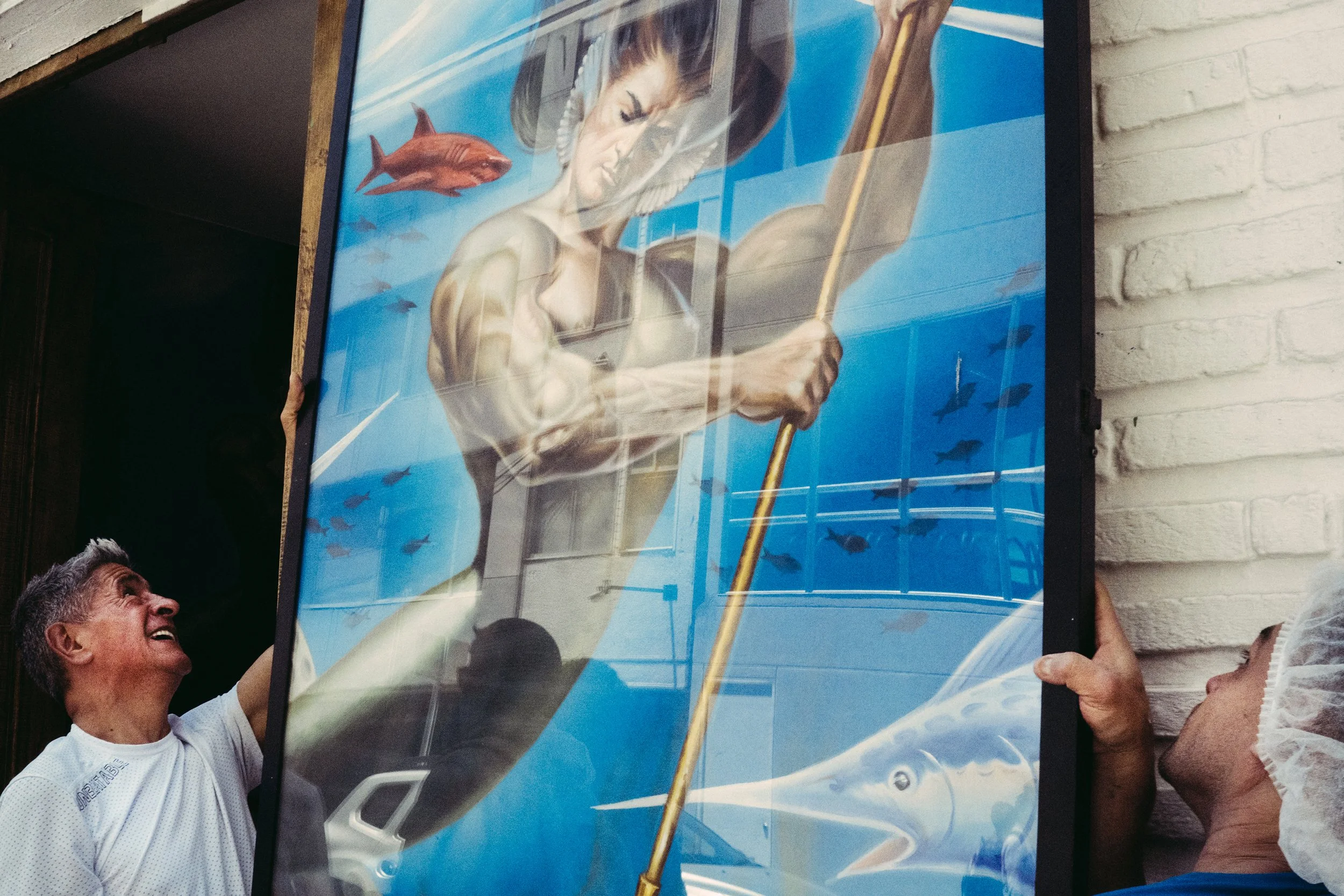 Hombre feliz sosteniendo un cartel con imagen de una figura espiritual con barba, barca y fondo marino con peces, en un entorno de reunión social.