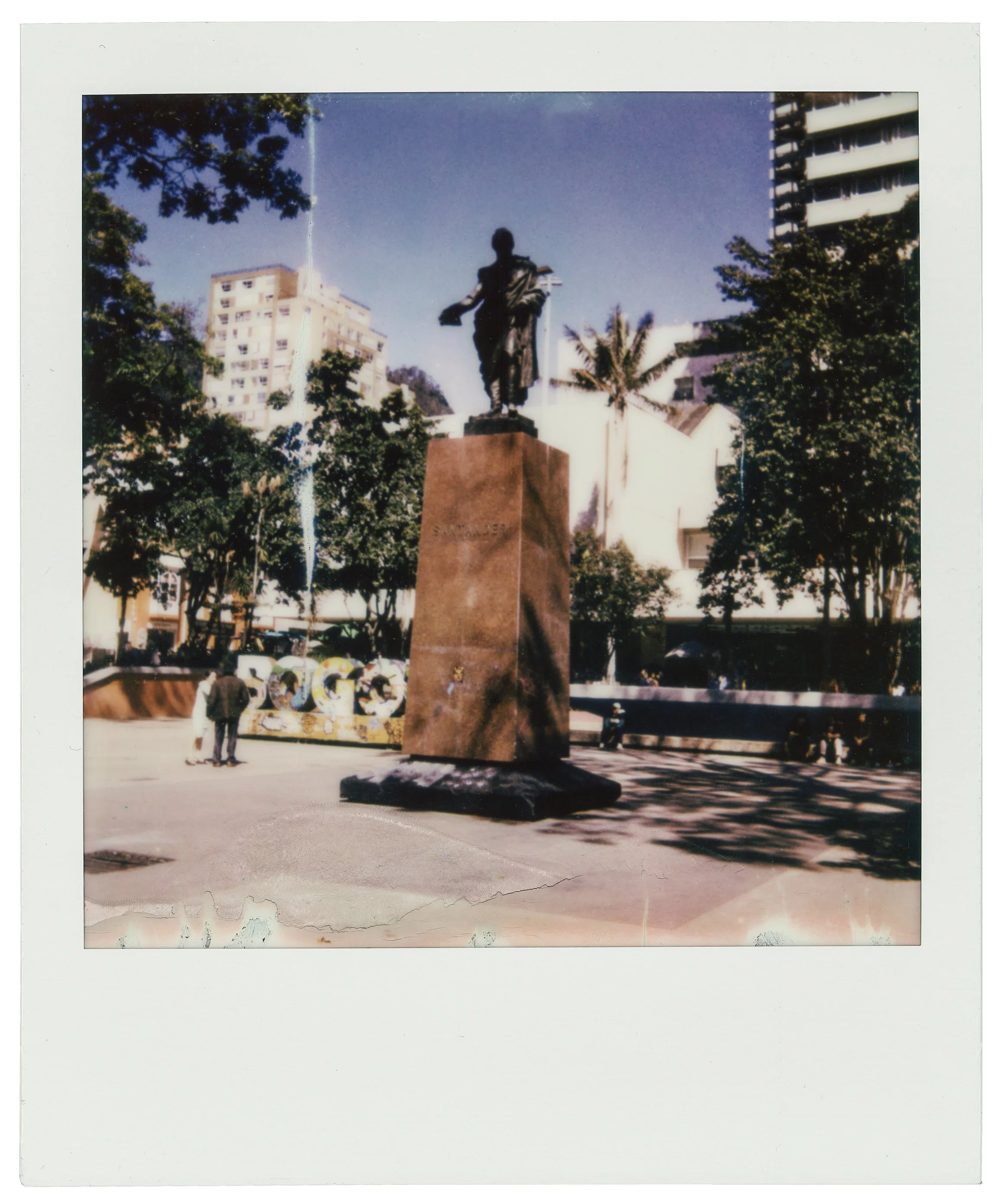 Escultura de una figura humana de bronce sobre un pedestal de piedra en medio de un parque con árboles y edificios modernos al fondo.