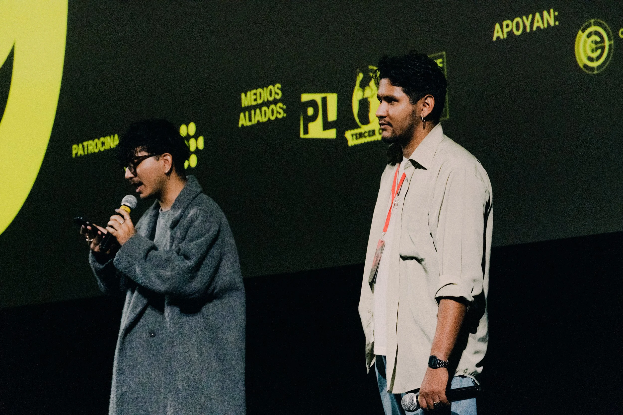 Dos personas hablando en un evento, una mujer con gafas y chaqueta gris, un hombre con camiseta blanca y accesorios, ambos sosteniendo micrófonos, fondo con logotipos y texto en español.