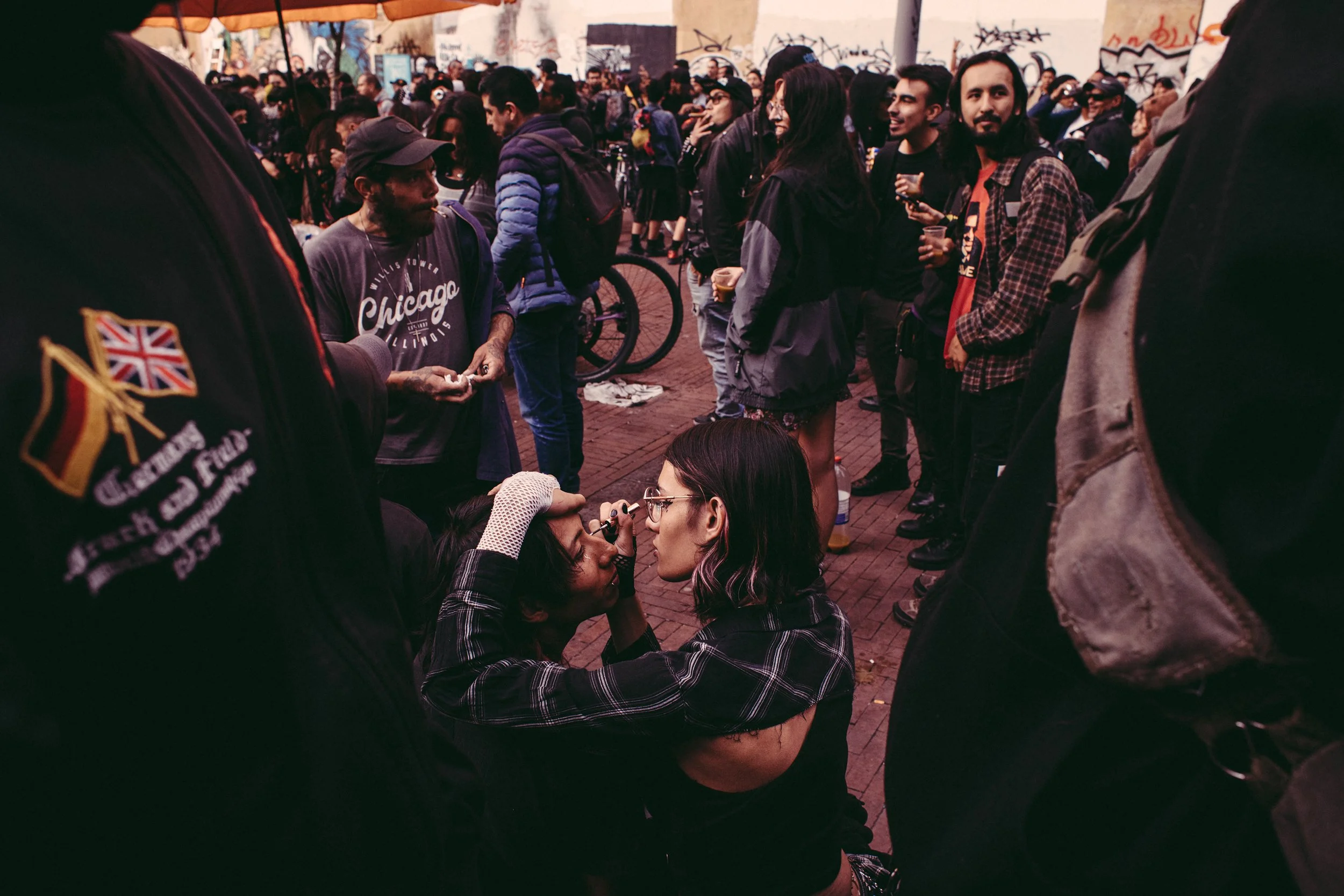 Muchísimas personas reunidas en una calle, con graffiti en las paredes de fondo, algunas conversando, algunas con bebidas en las manos, un ambiente urbano y social.