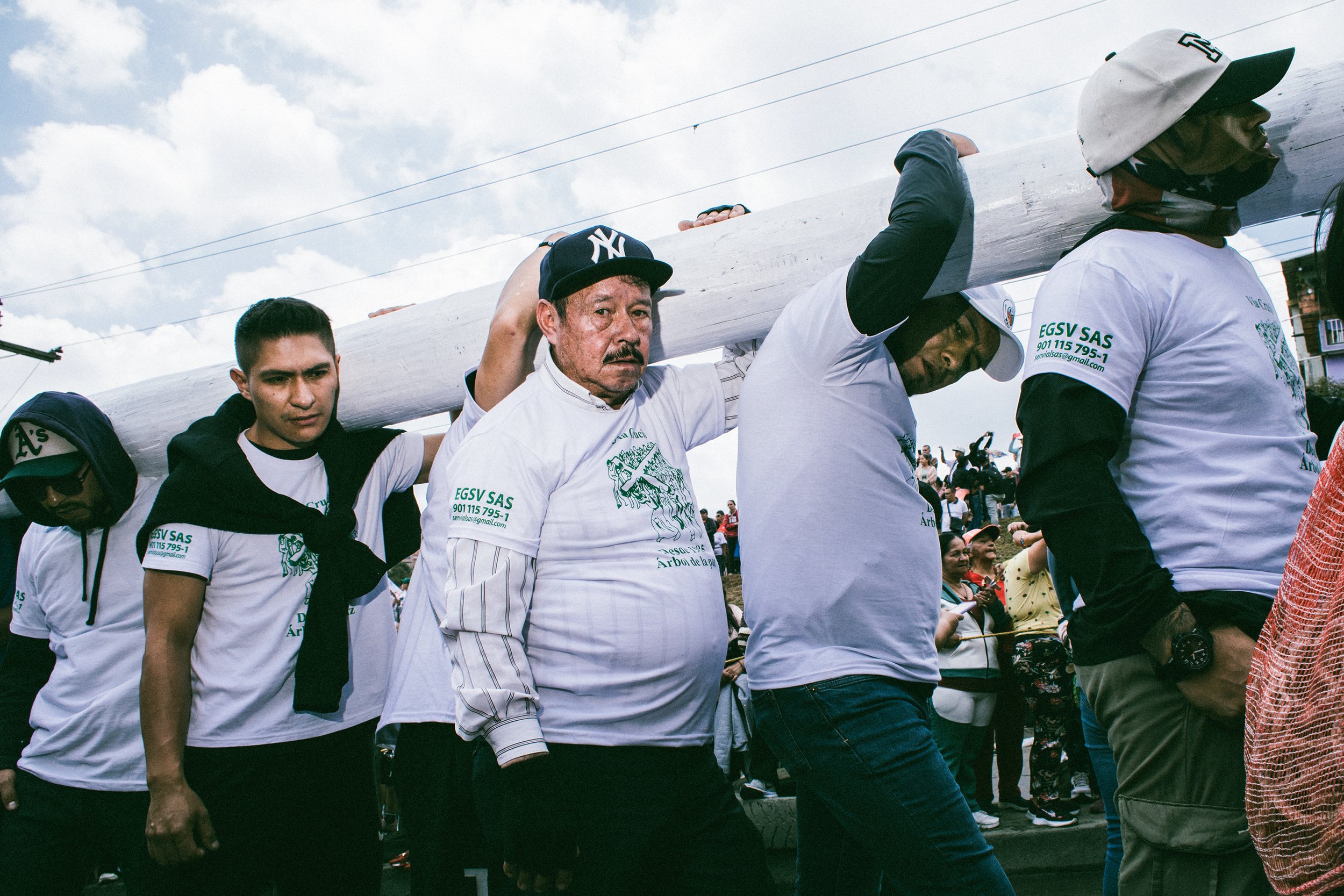 Personas participando en una marcha, algunas llevan una cruz grande sobre sus hombros, en un evento al aire libre, rodeados de muchas otras personas.