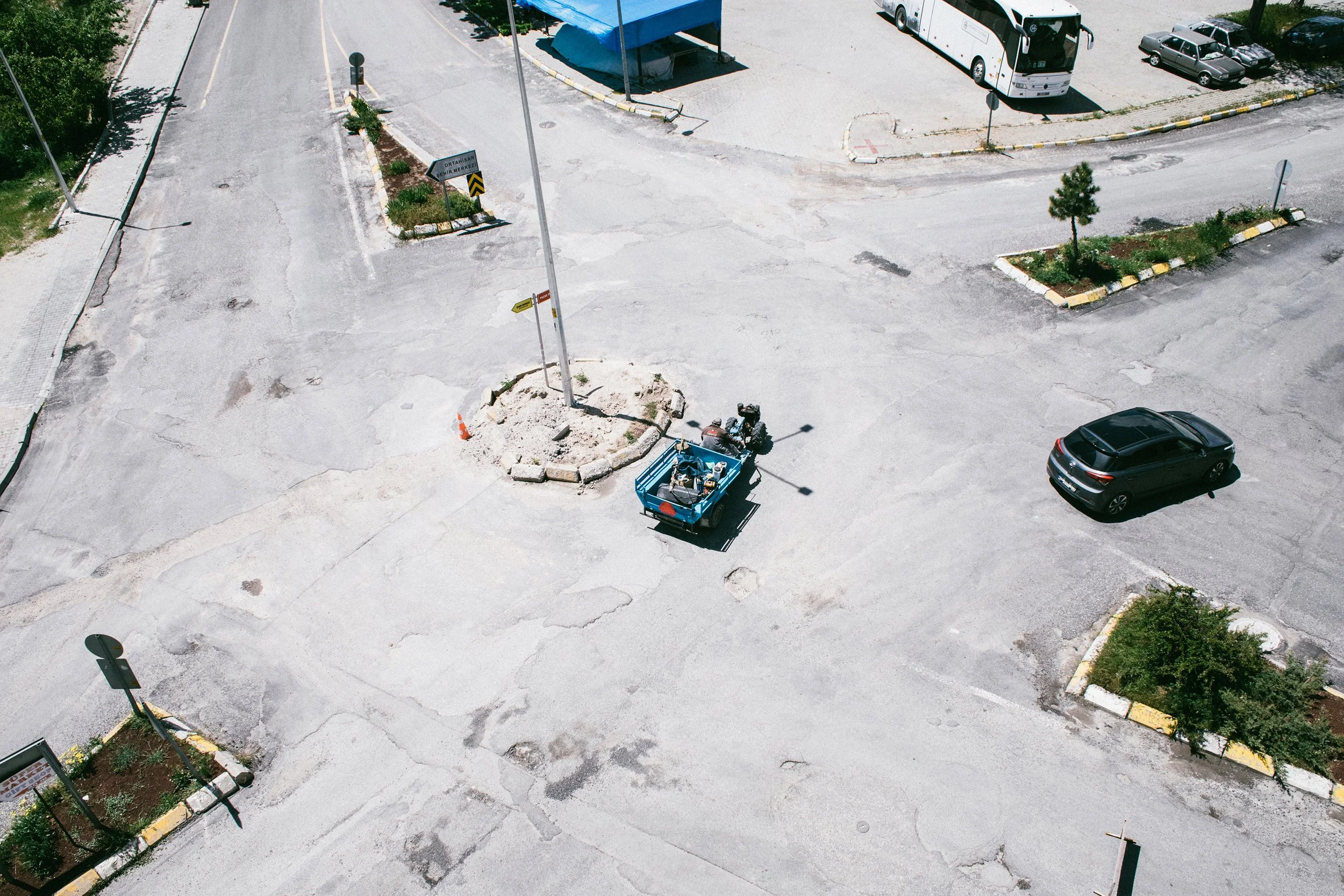 Estacionamiento con varias áreas delimitadas, algunos árboles, una camioneta azul en reparación, un autobús, varios vehículos, señalización vial y puntos de sombra.