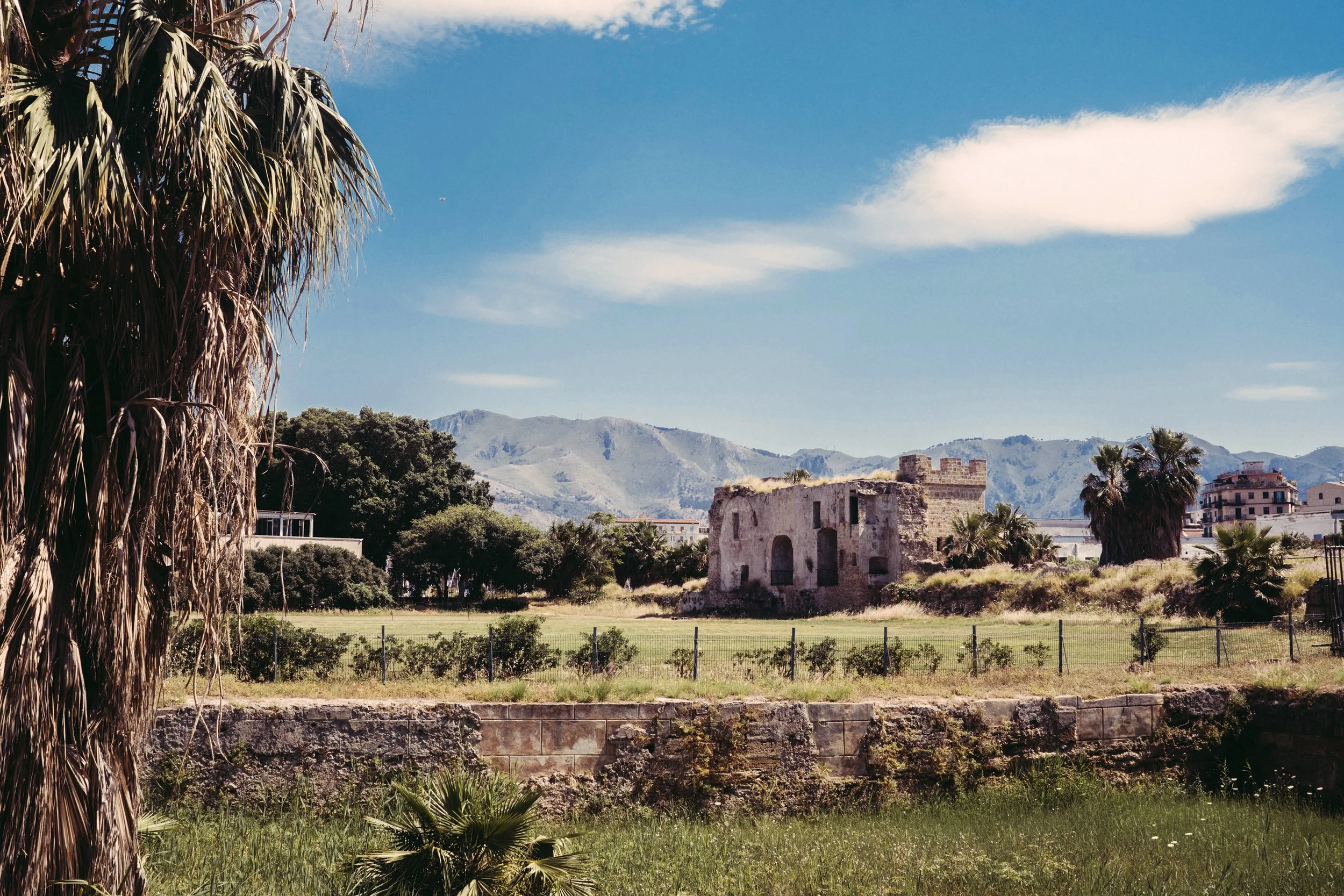 Paisaje con árboles, ruinas antiguas y montañas al fondo bajo un cielo azul con algunas nubes.
