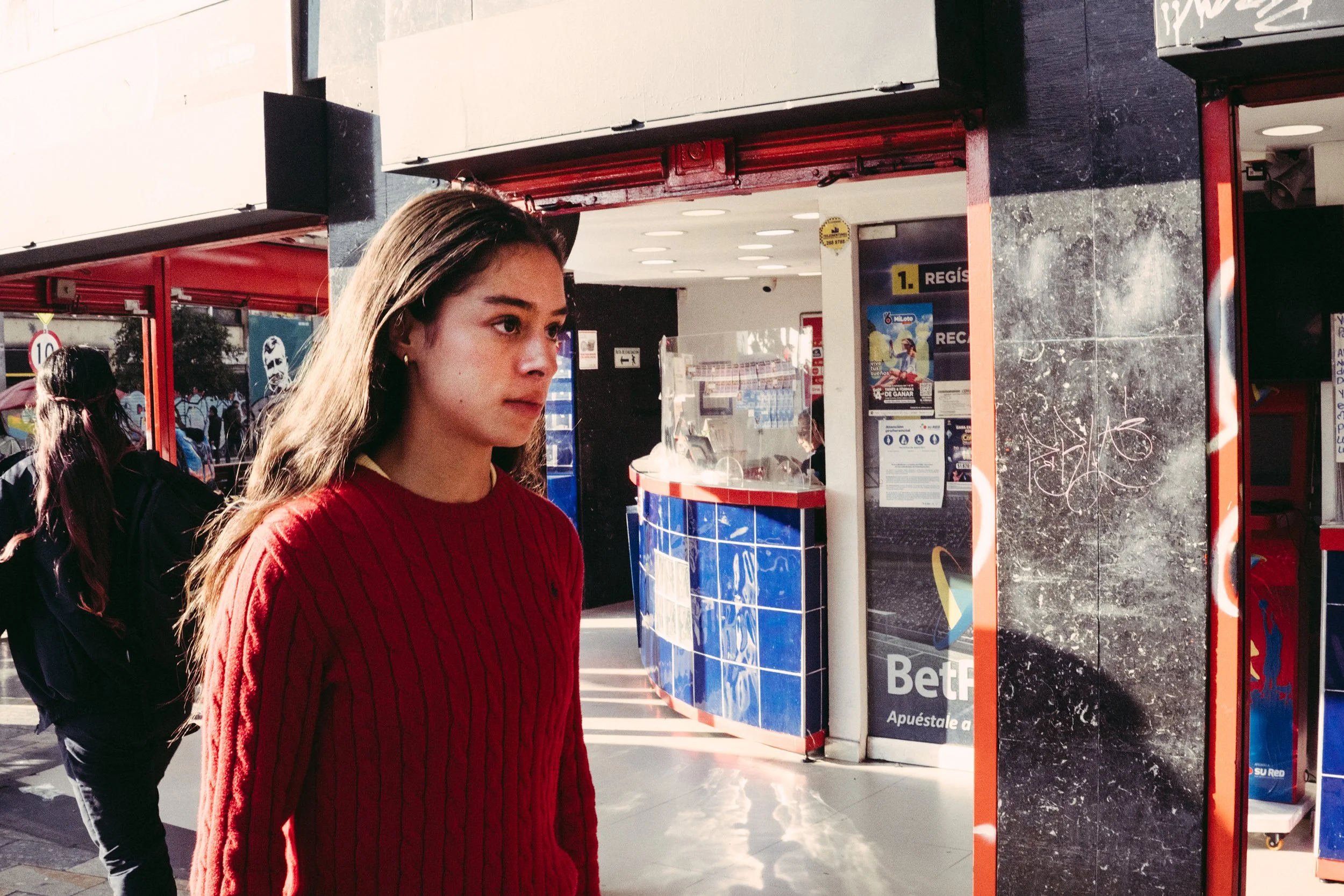 Joven con cabello largo y rubio, usando una sudadera roja, de pie en una estación de autobuses o metro, con fondo de kiosco y señalización en español.