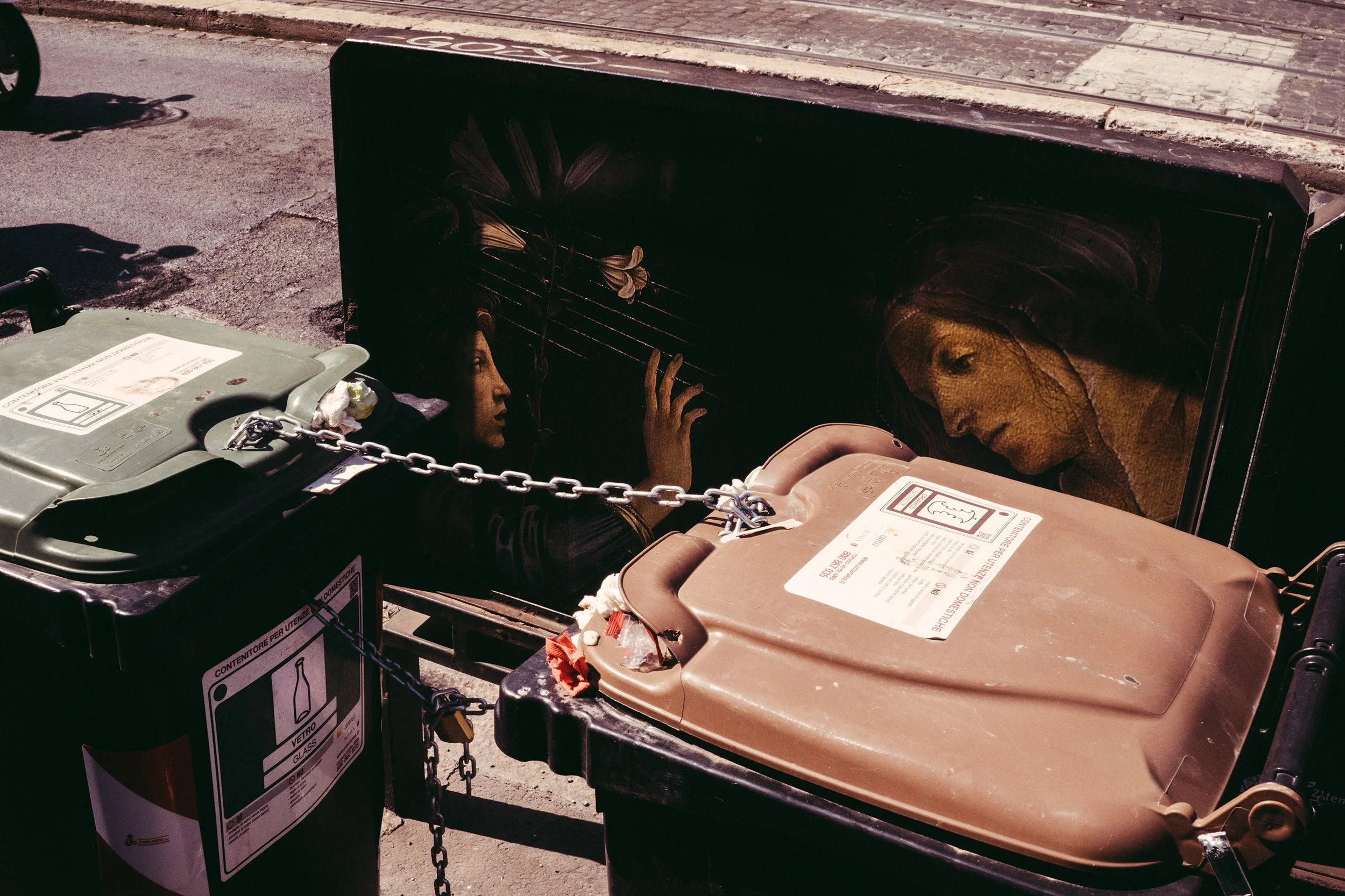 Dos contenedores de basura, uno negro y otro marrón, con etiquetas y cadenas cierres, en una calle pavimentada. En el fondo, una acera y una vía con rieles del tren.
