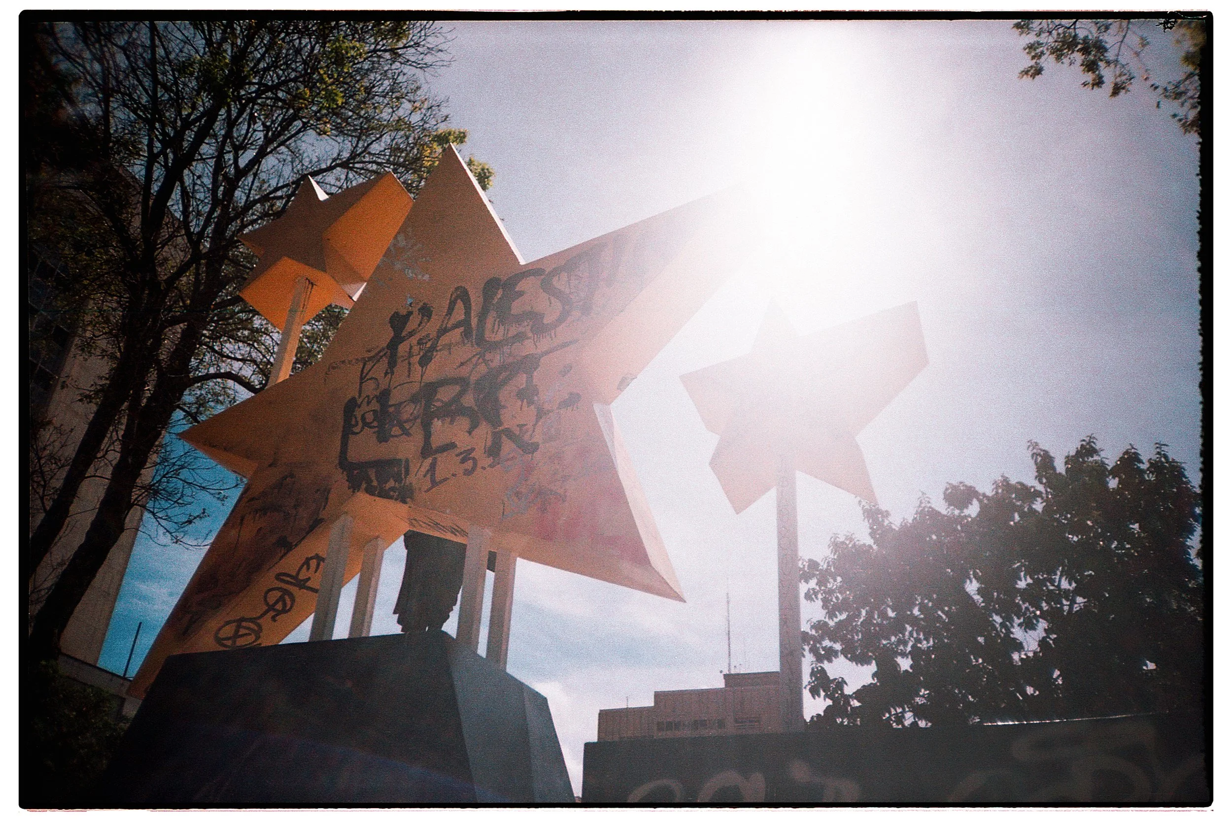 Escultura pública con letras y gráficos, situada en un parque con árboles y edificios al fondo, vista desde abajo con el sol brillante