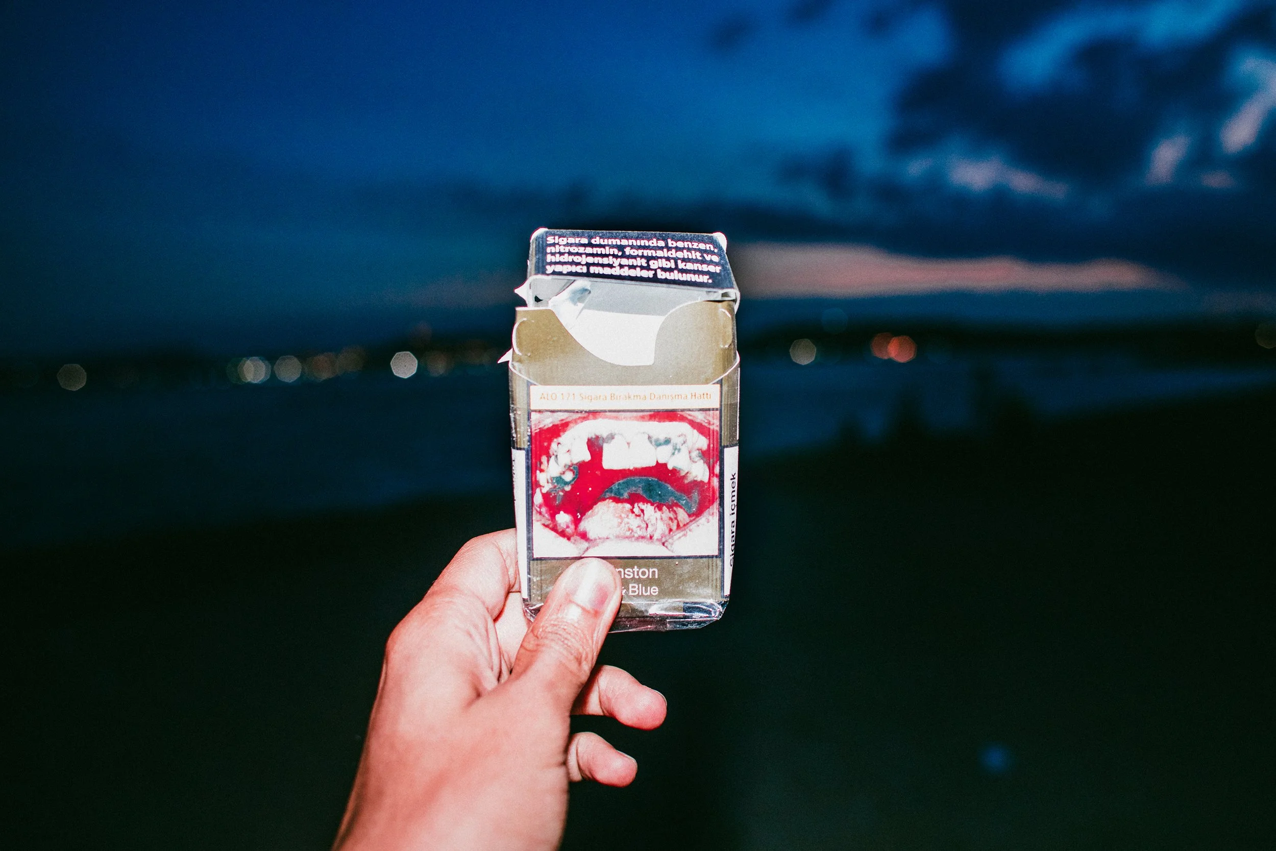 Una mano sostiene un paquete de cigarrillos con el fondo de un cielo nublado y la ciudad a distancia.