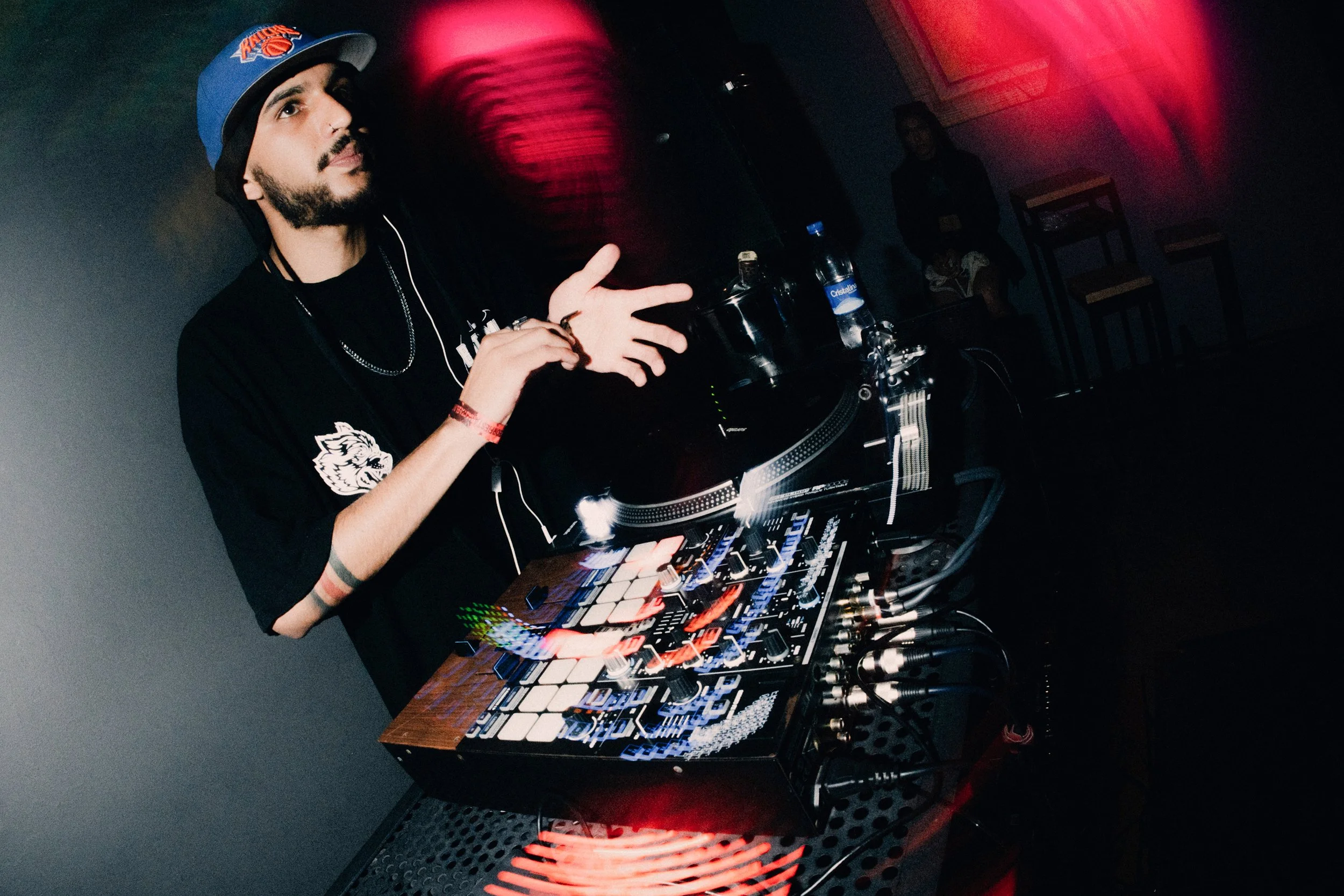 Un DJ con barba, sombreado por un gorro de la dijo Knicks, mezclando música en una cabina con varios equipos digitales y plato giratorio, en un ambiente oscuro con luces rojas y verdes.