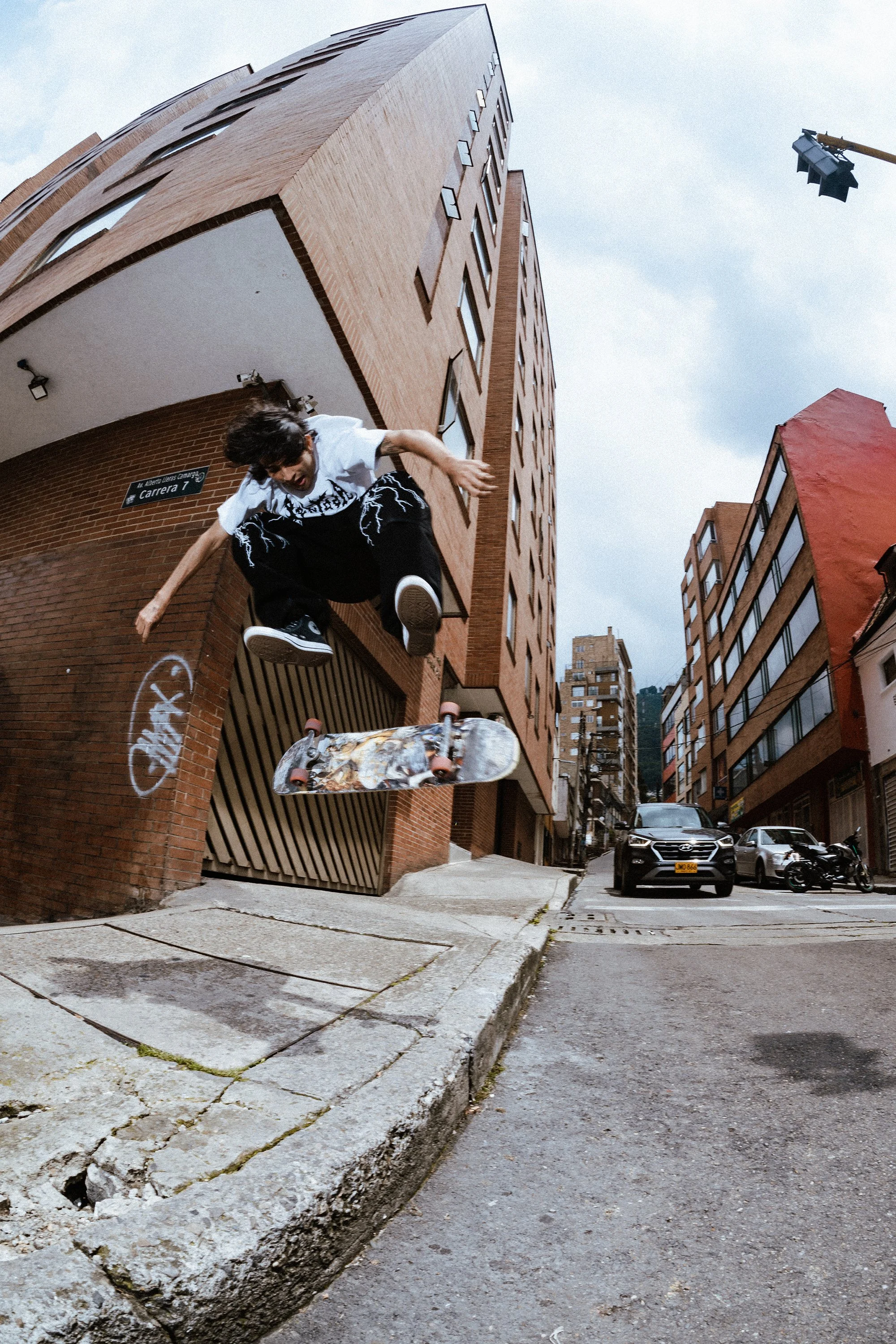 Un joven haciendo un truco en una patineta en la calle de una zona urbana con edificios de ladrillo y carros estacionados, durante el día.
