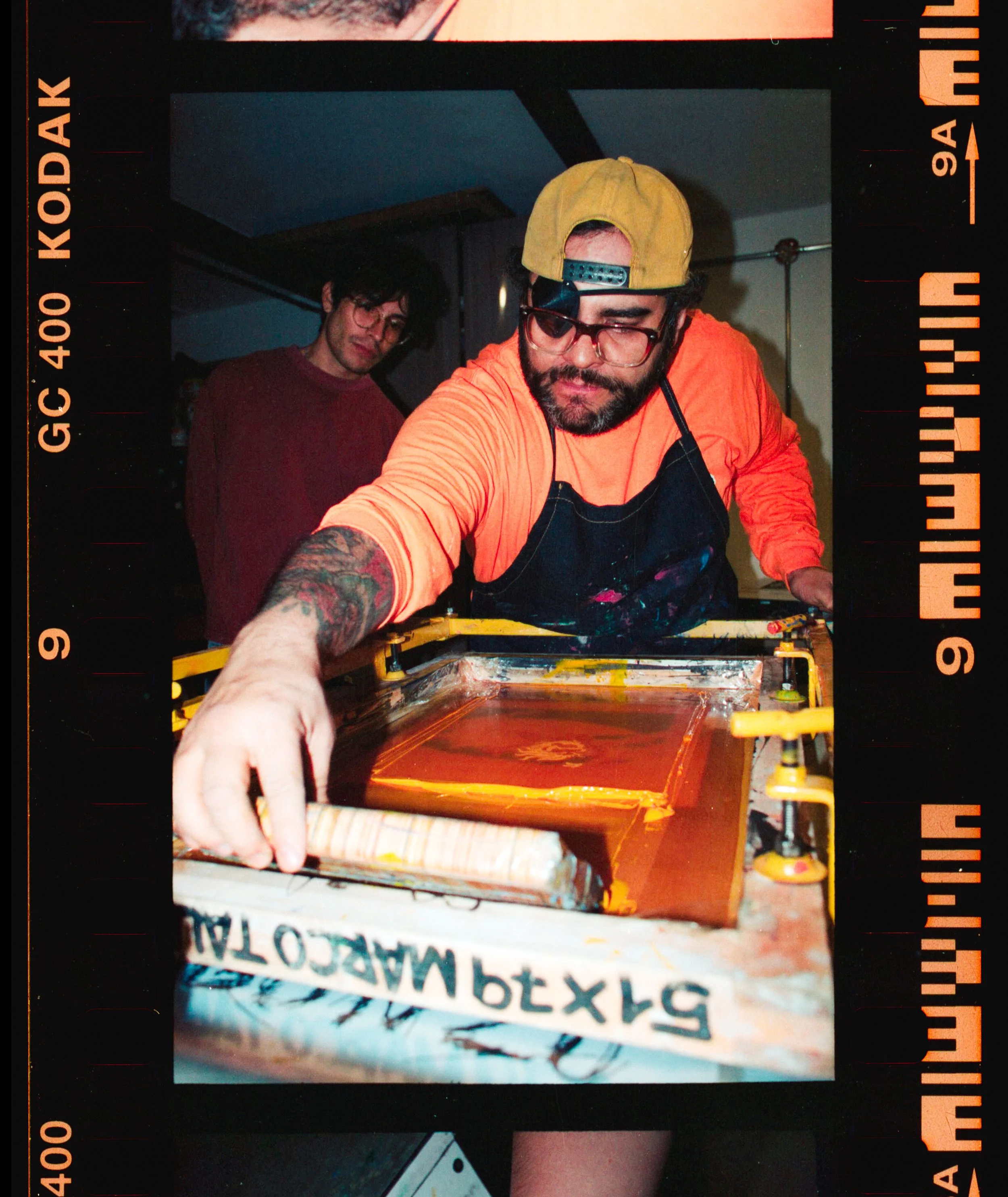 Dos personas en un taller, una de ellas con gafas y gorra amarilla, trabajando en un proyecto de impresión en cama plana con tinta naranja.