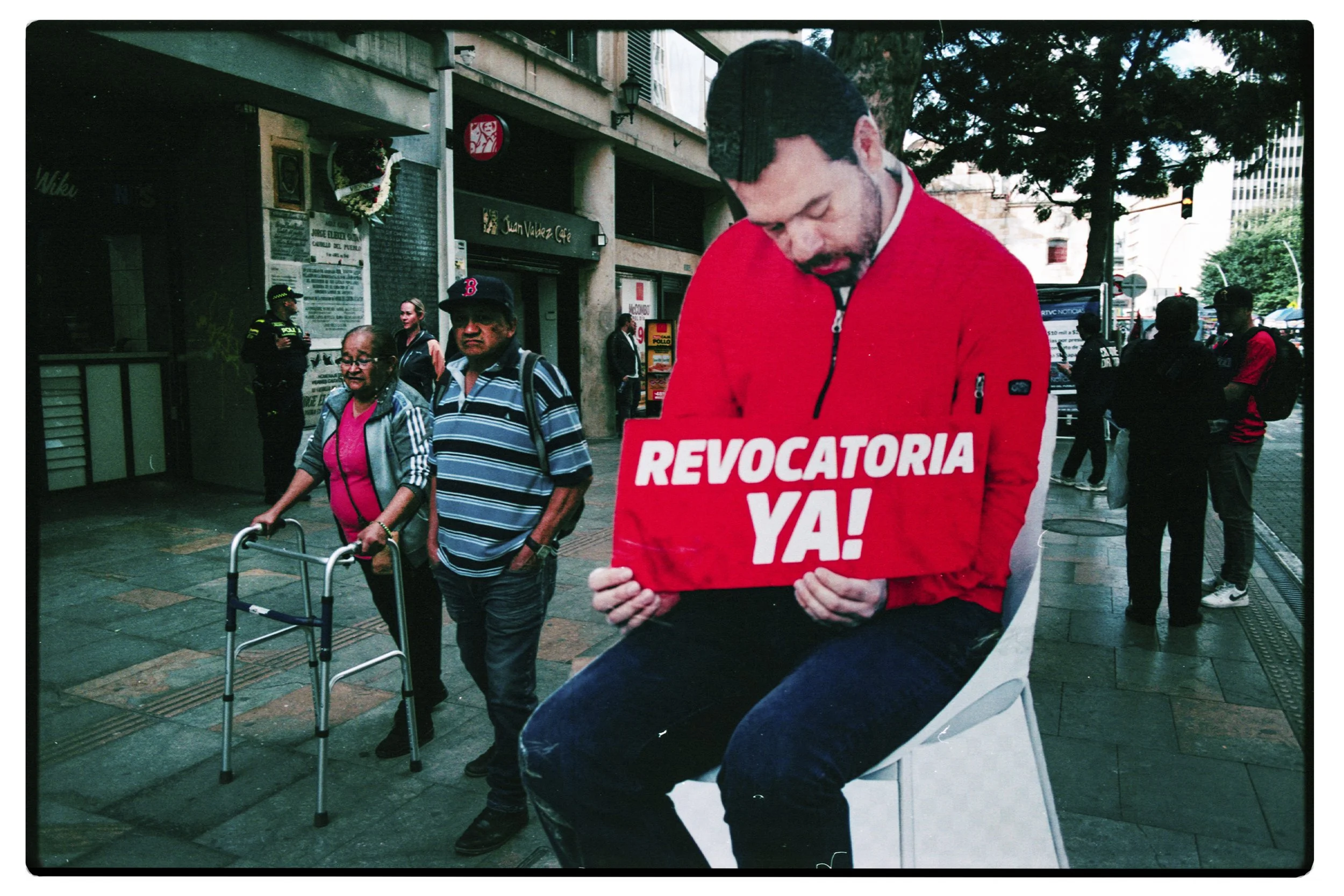 Manifestante con camiseta roja que sostiene cartel con la palabra 'REVOCATORIA YA' en una calle concurrida, alrededor de varias personas y policías.