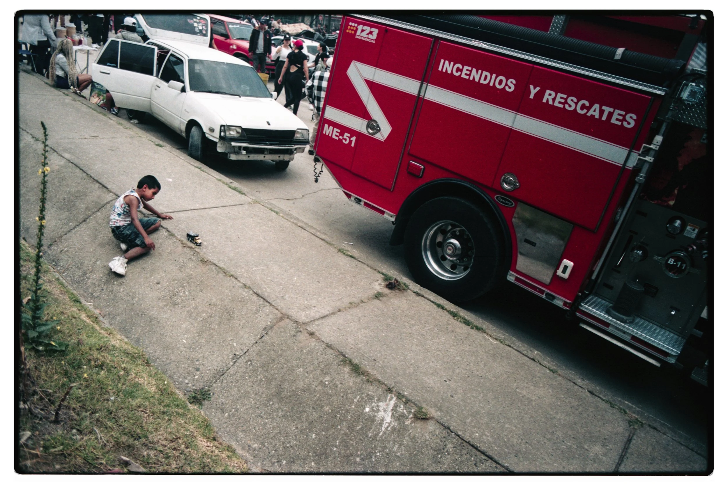 Una escena en la calle donde hay una emergencia con un auto blanco que aparentemente sufrió un accidente, un camión de bomberos rojo con la leyenda 'Incendios y Rescates', varias personas en el fondo y una niña jugando con un carrito en la acera.