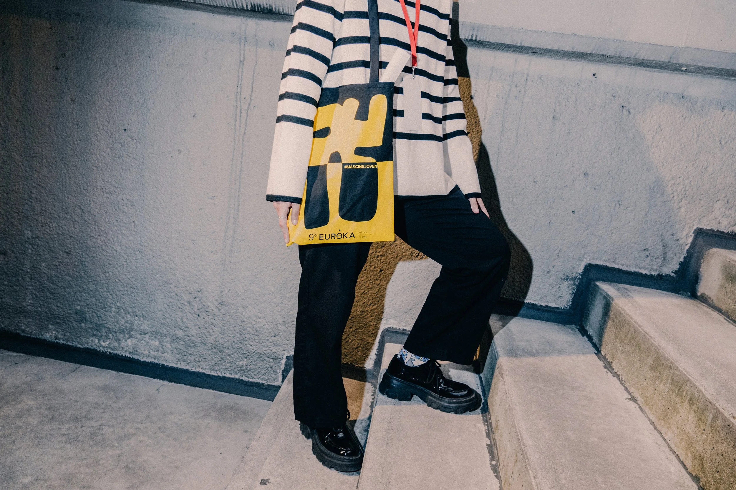 Persona sosteniendo una bolsa de color amarillo y negro con texto y hashtags, vestida con suéter de rayas negras y blancas, pantalones negros y botas negras, en una escalera de concreto con paredes grises.