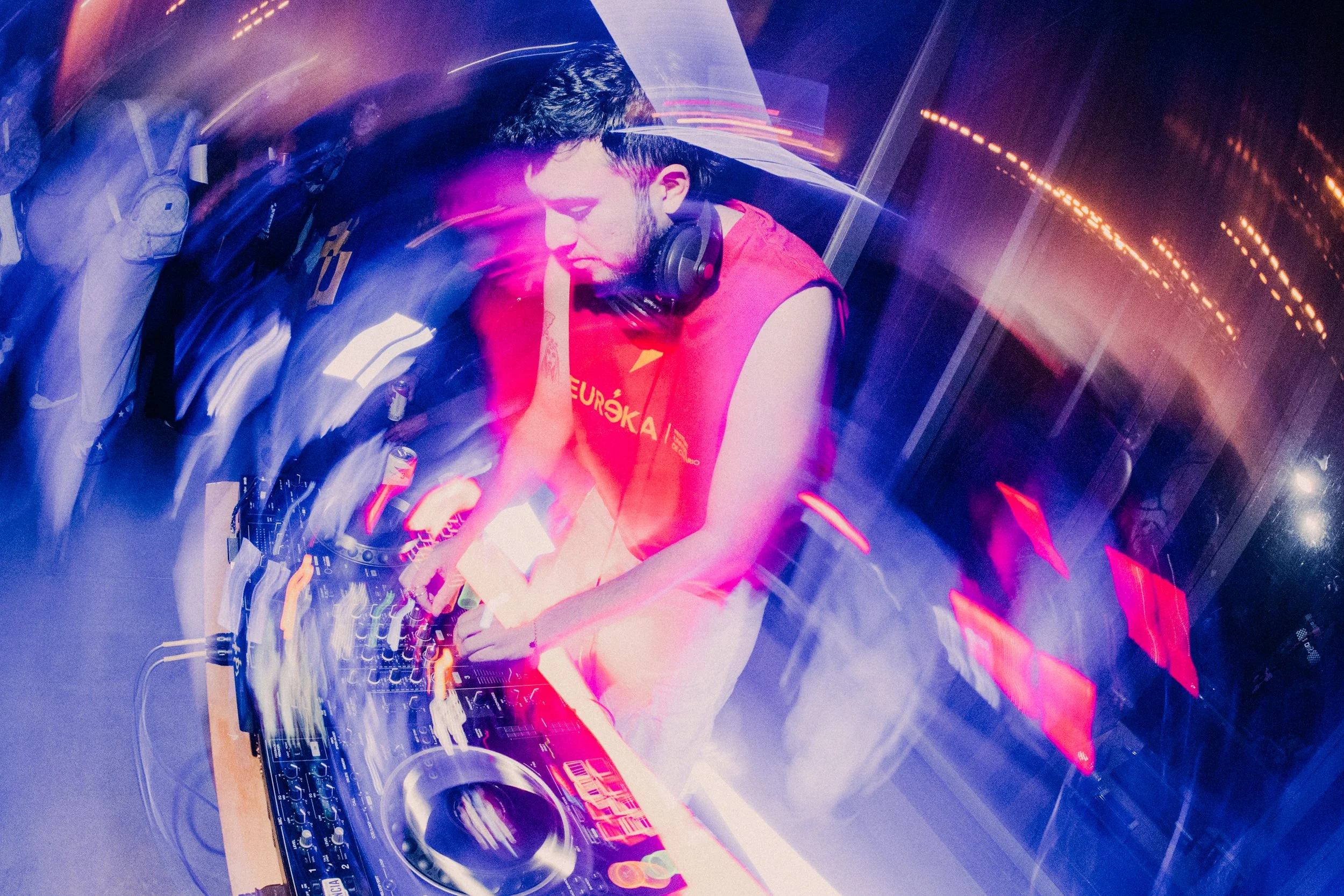 Un DJ con auriculares, mezclando música en un equipo de sonido en una cabina. Luces de colores y efectos de movimiento en el entorno.