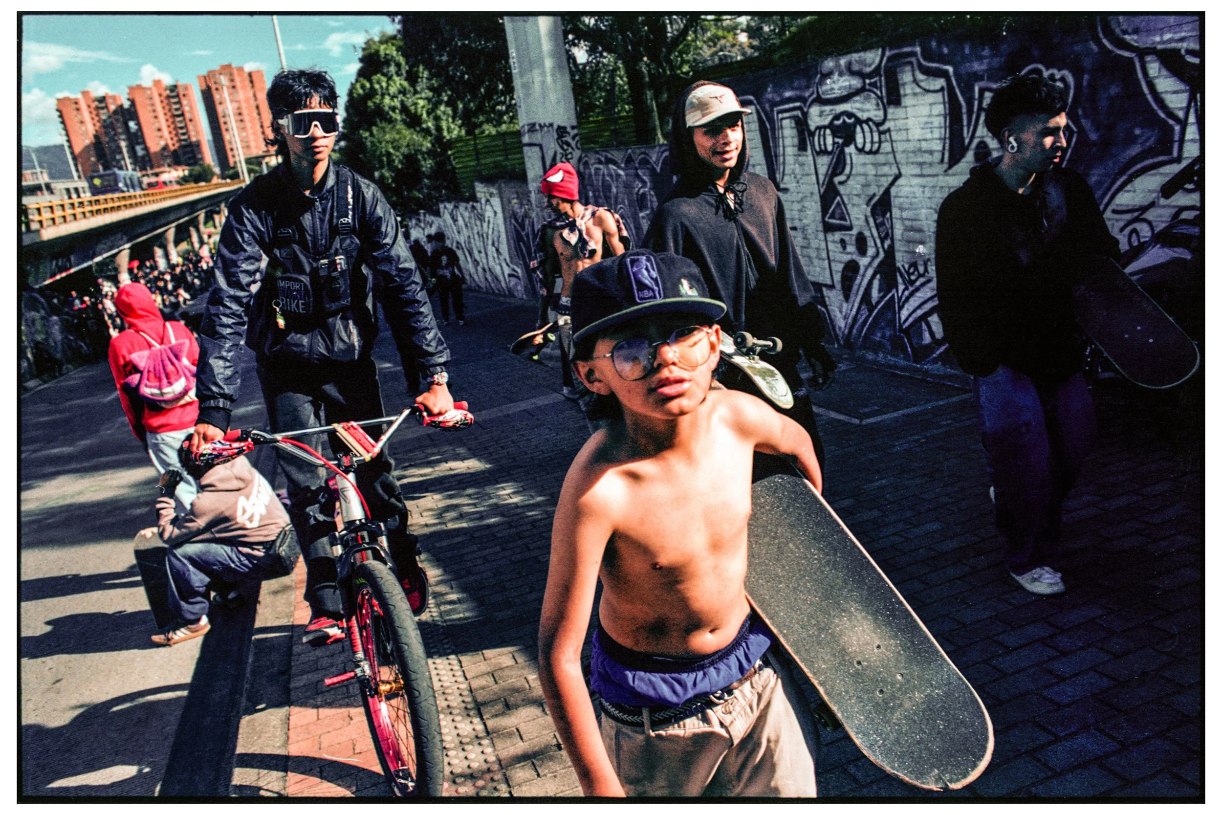 Joven sin camiseta con gafas con un skate, rodeado de personas en un mural callejero.