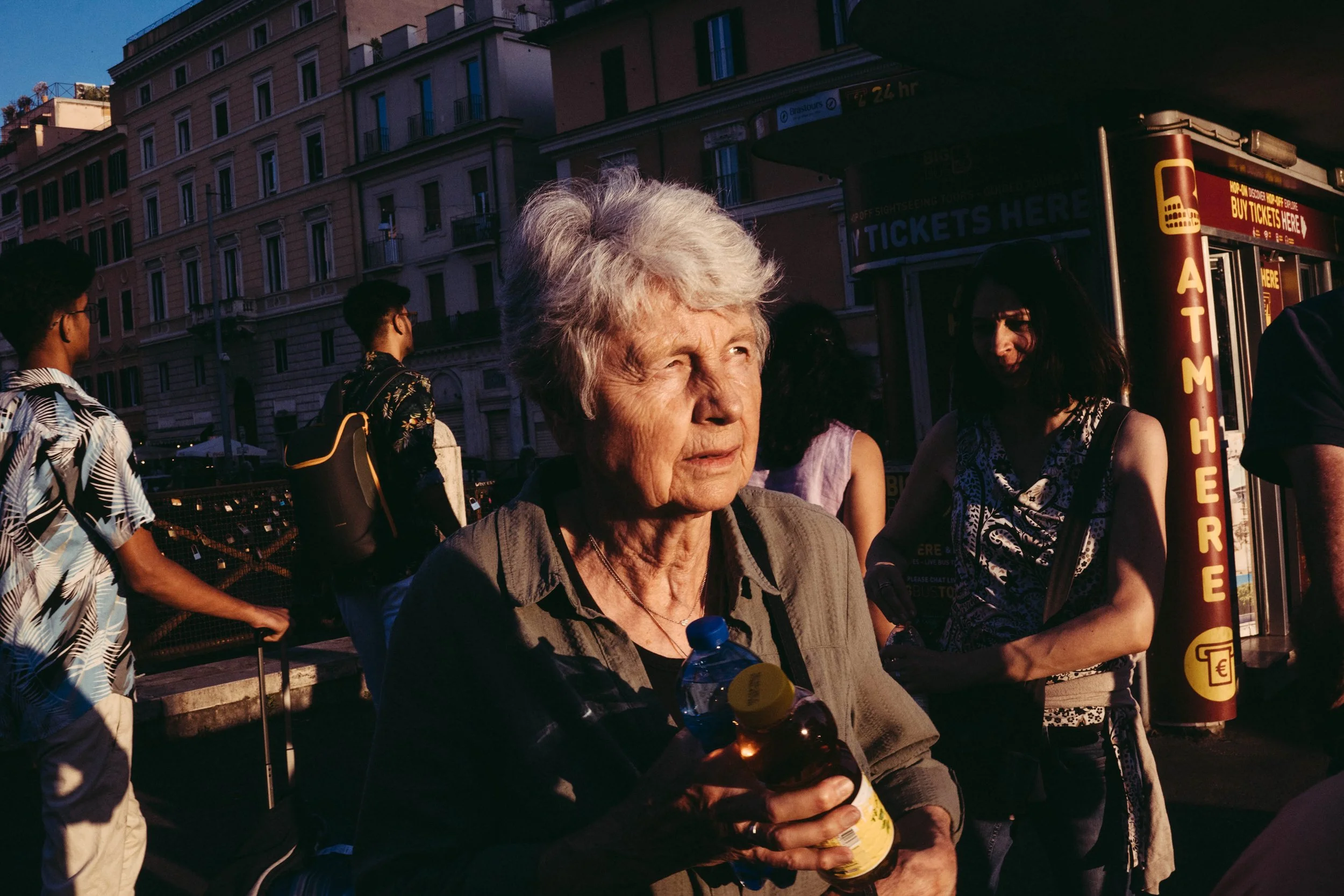 Una mujer de cabello gris con expresión pensativa, sosteniendo una botella y una cuchara, rodeada de varias personas en un espacio al aire libre en una ciudad, con edificios y un puesto de boletos en el fondo.