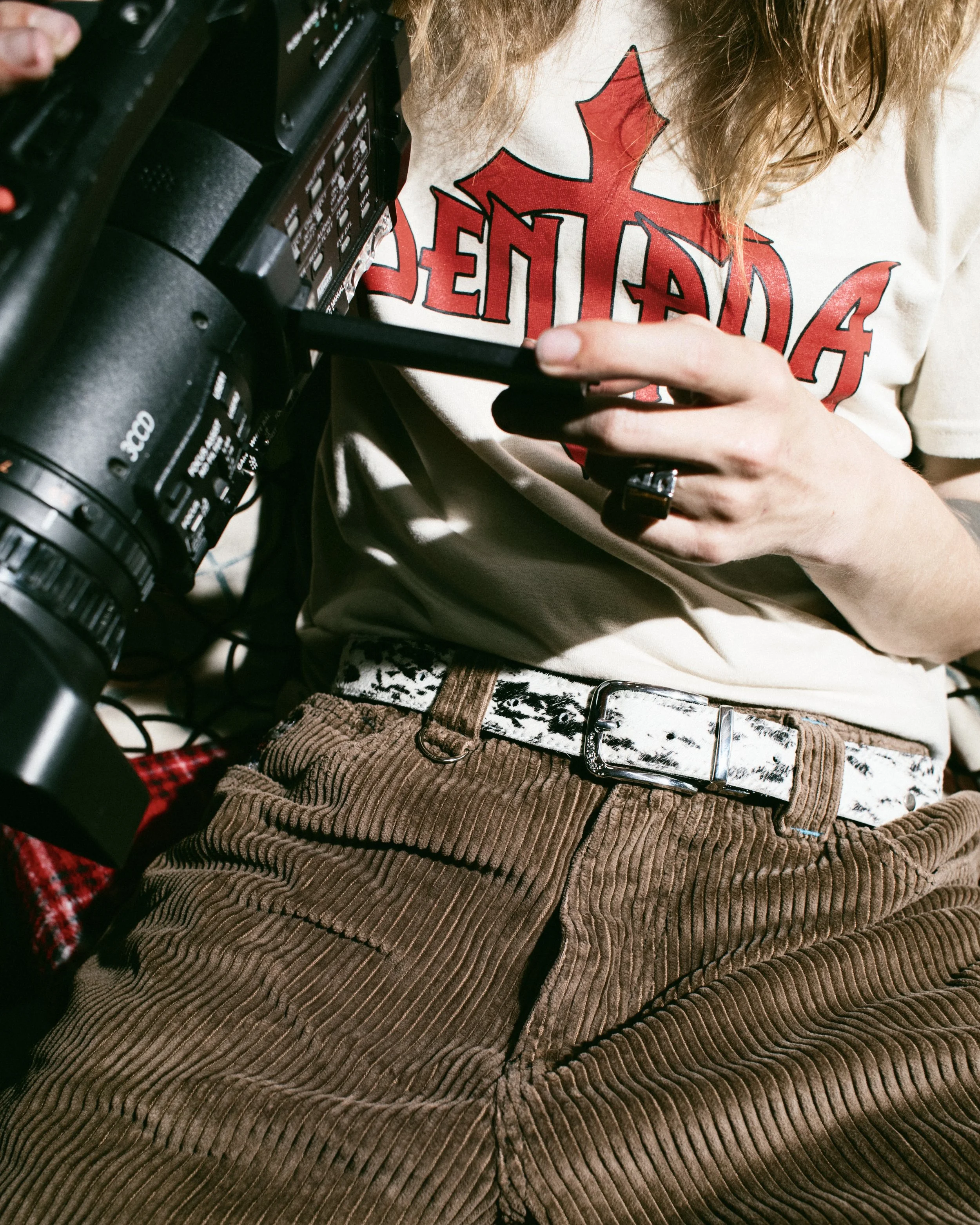 Persona sujetando una cámara de video o fotografía, vistiendo una camiseta blanca con un símbolo de una cruz roja y letras en negro. Usa pantalones de mezclilla color marrón con cinturón de tela con estampado, y tiene cabello rubio y largo.