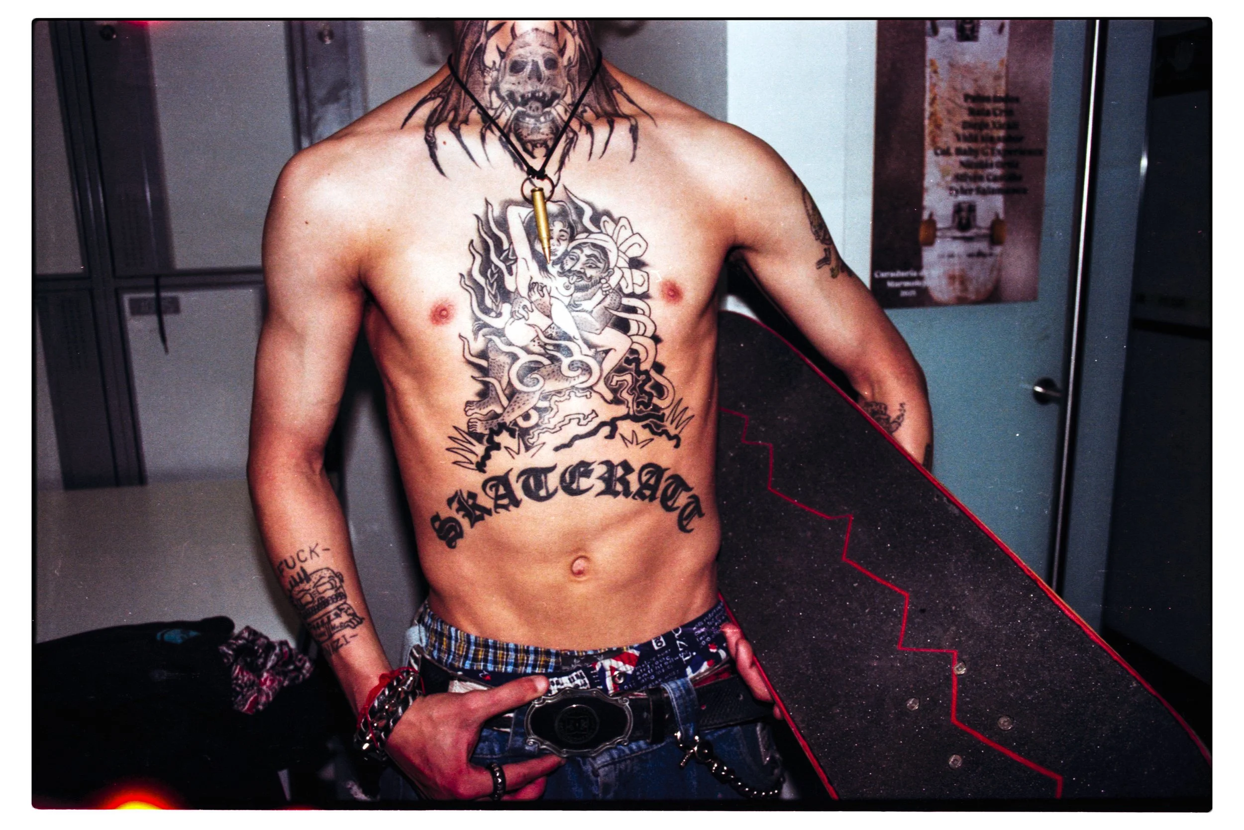 Hombre con tatuajes en el torso, usando pantalones cortos de cuadros y varias pulseras, tomando una selfie en un espejo en un vestuario