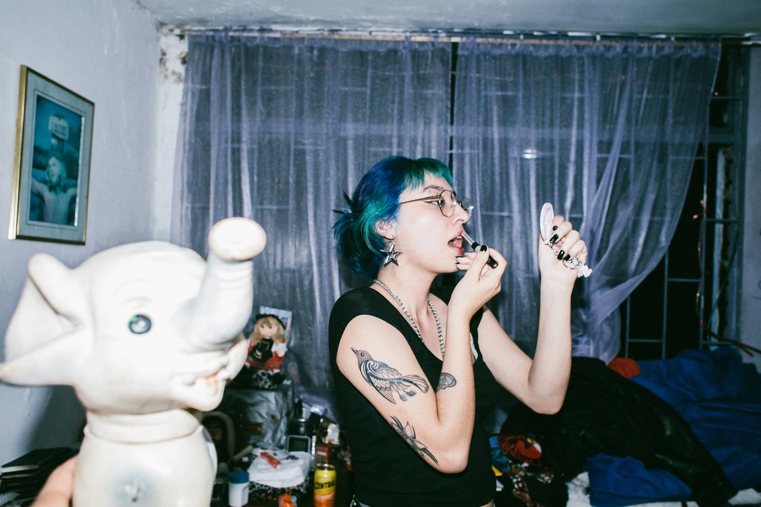 Una mujer con cabello teñido de azul y verde, usando gafas y viendo en un espejo mientras se aplica maquillaje. Tiene tatuajes en el brazo y usa pendientes de estrella. En primer plano, una escultura de Cerdo de porcelana y en el fondo, una cortina d