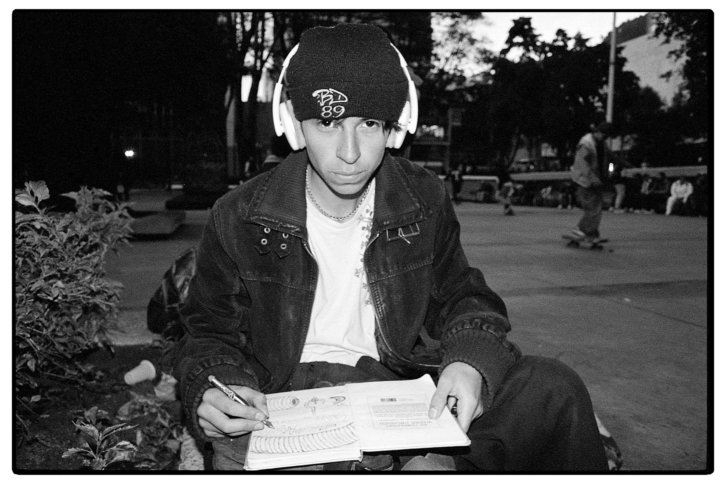 Joven sentado en un parque, con gorra y auriculares, escribiendo en un cuaderno.