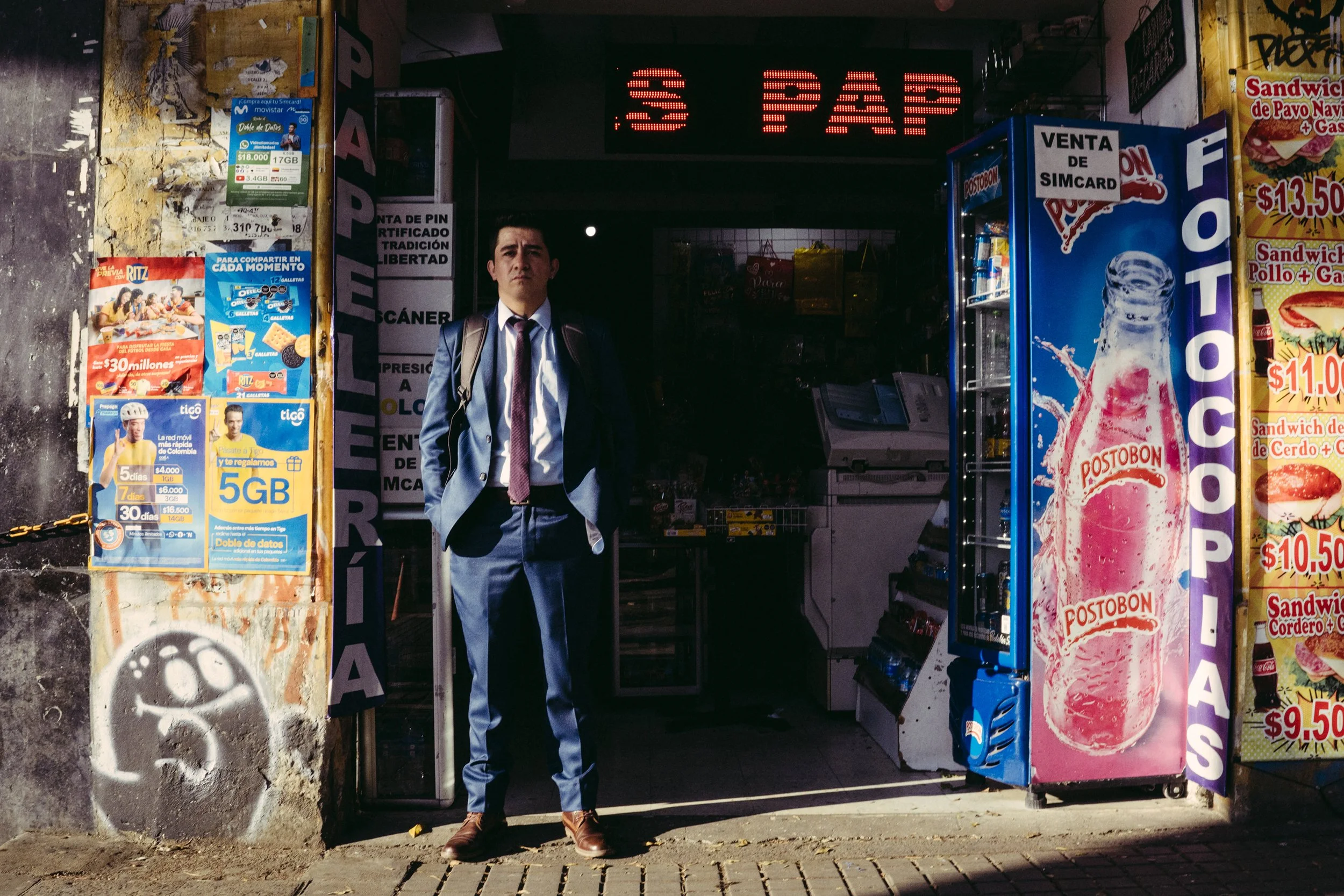 Hombre vestido con traje y maletín, de pie frente a una tienda de conveniencia con anuncios de tarjetas SIM y productos en vitrinas, en un entorno urbano.