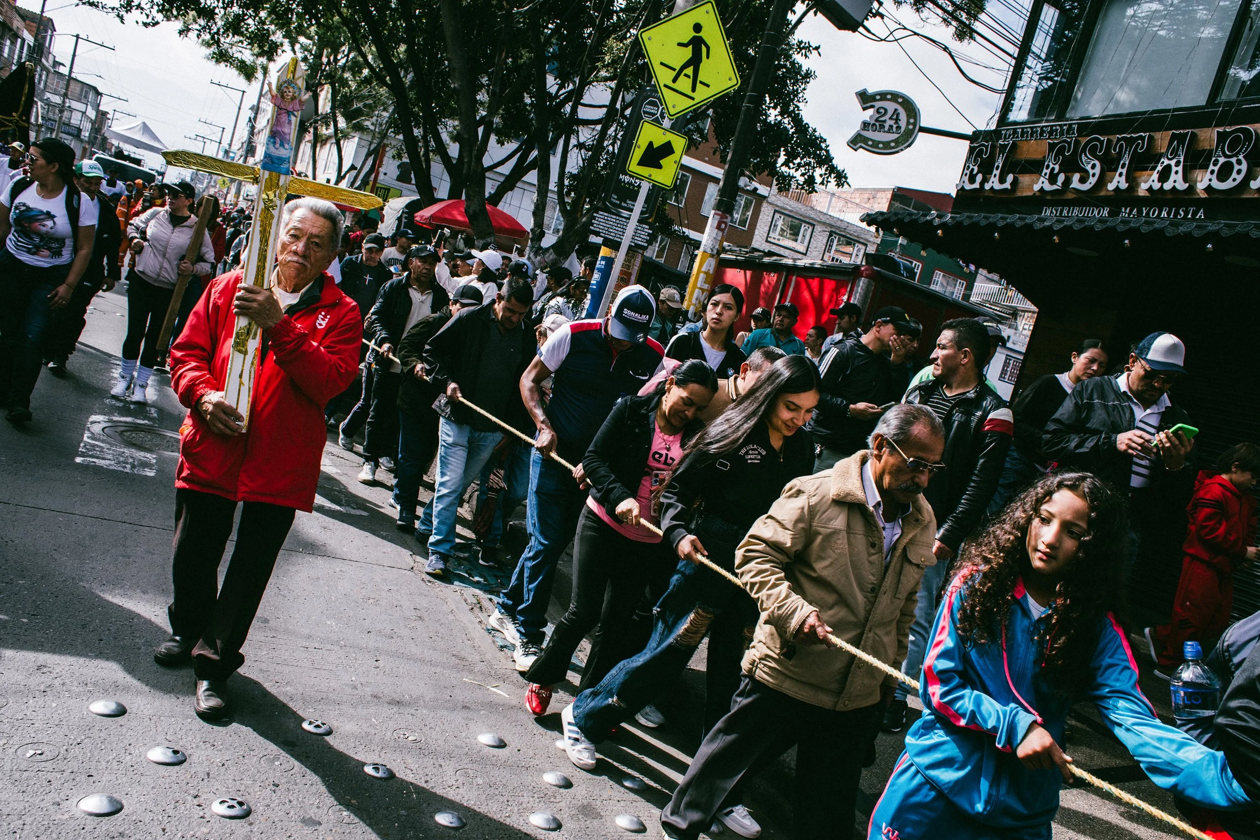 Grupo de personas participando en una procesión o evento tradicional en la calle, con alguien sosteniendo una figura religiosa y varias personas uniéndose a una cuerda, en un ambiente urbano con señales de tránsito y tiendas.
