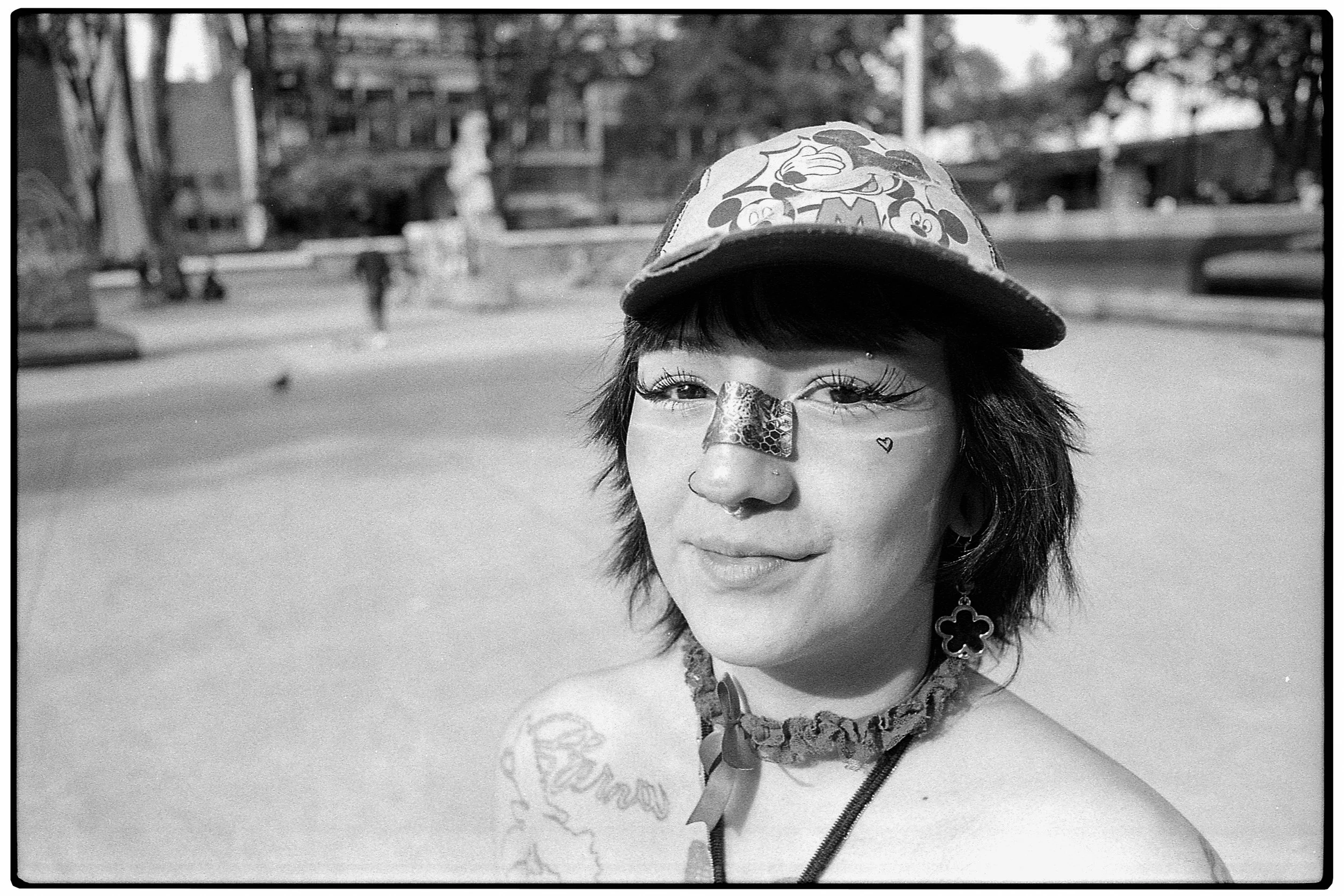 Mujer joven con sombrero decorado con personajes de dibujos animados, con accesorios como pendientes con forma de flor, en un parque, sonrisa casual en rostro, en blanco y negro.