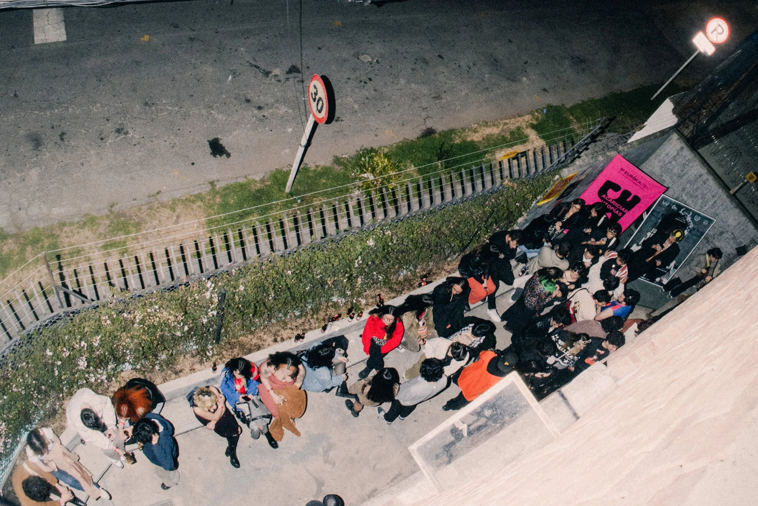 Personas esperando en una fila en la acera de una calle en la noche, con tráfico vehicular visible en el fondo y señalizaciones de tránsito.