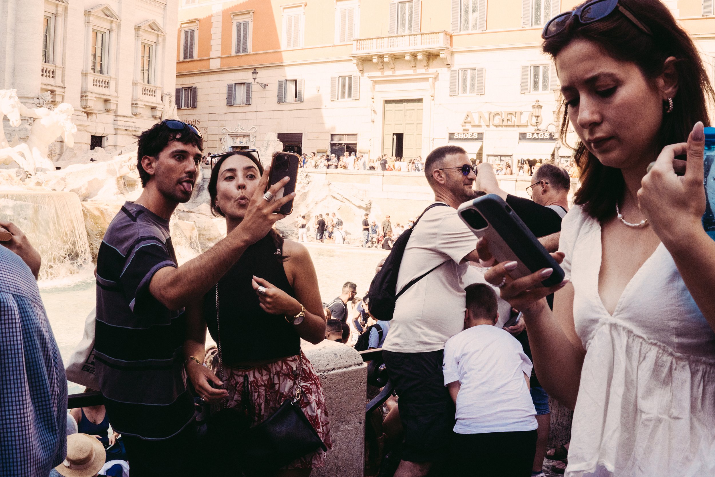 Grupo de personas en un lugar público, algunas usando gafas de sol, con edificios y una fuente en el fondo, algunos de ellos tomando fotos o usando sus teléfonos móviles.
